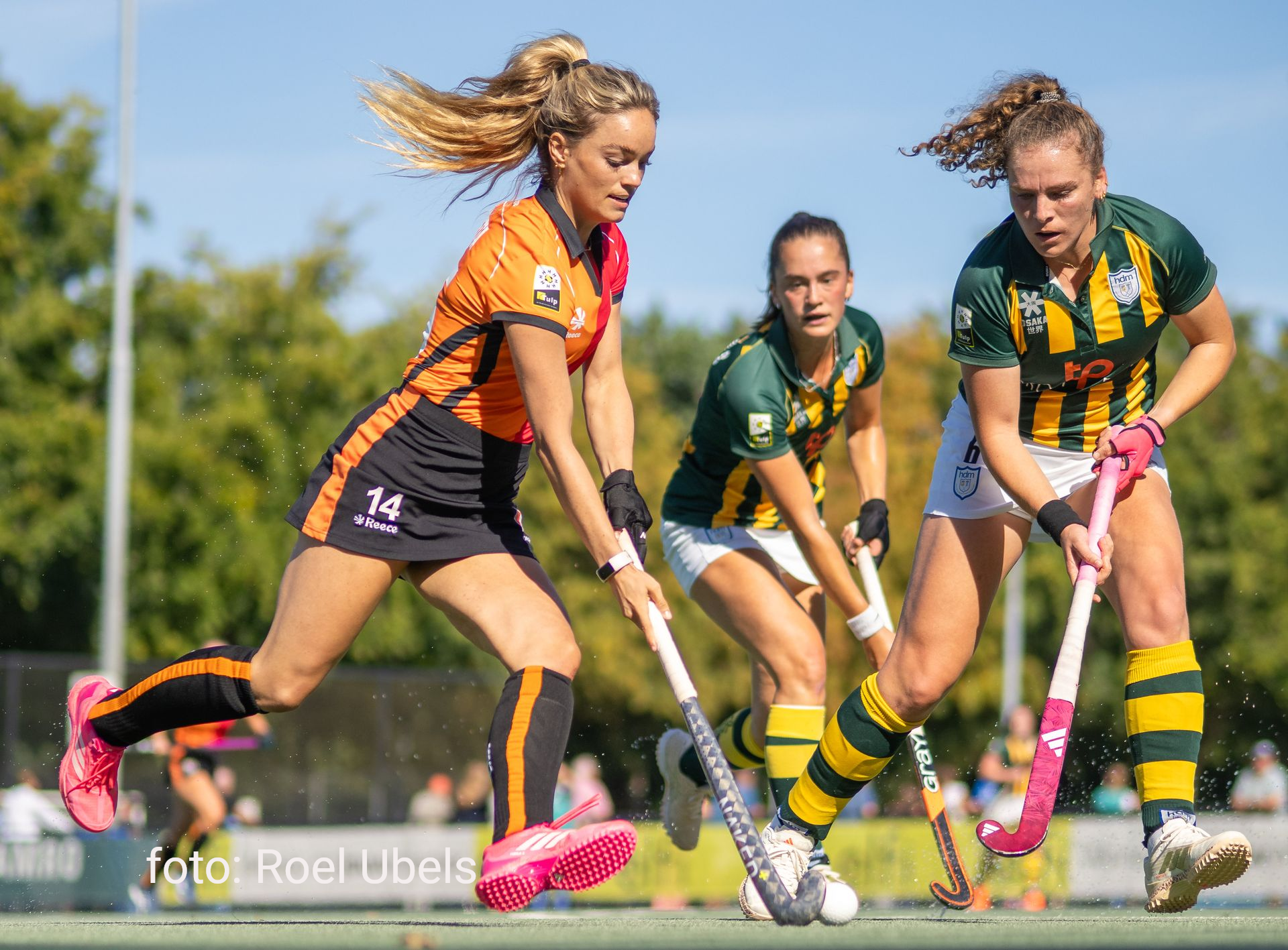 Hockeyspelers in actie, één in oranje, twee in groen, met sticks en bal.