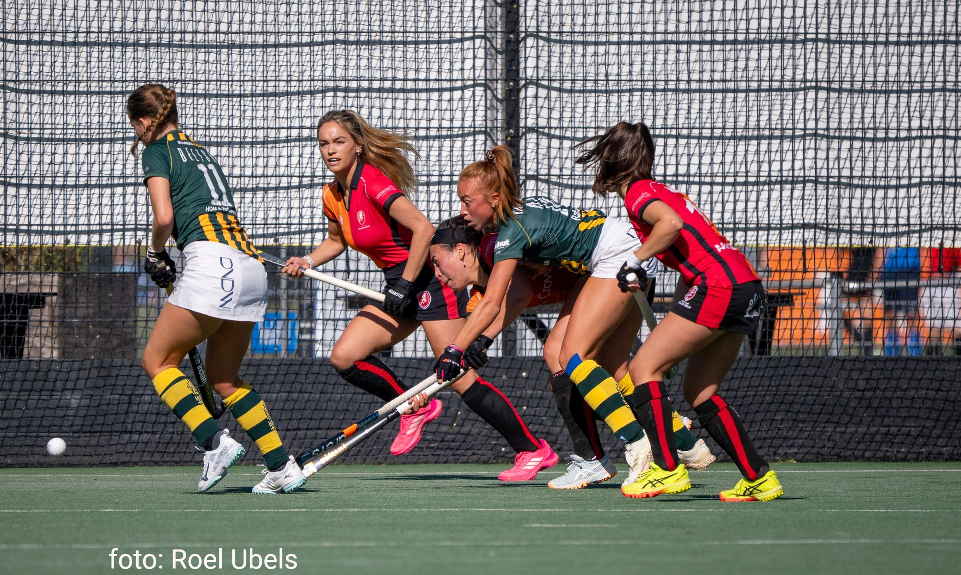 Hockeyspelers in actie, groene en rode tenues, bal op de grond, hek op de achtergrond.