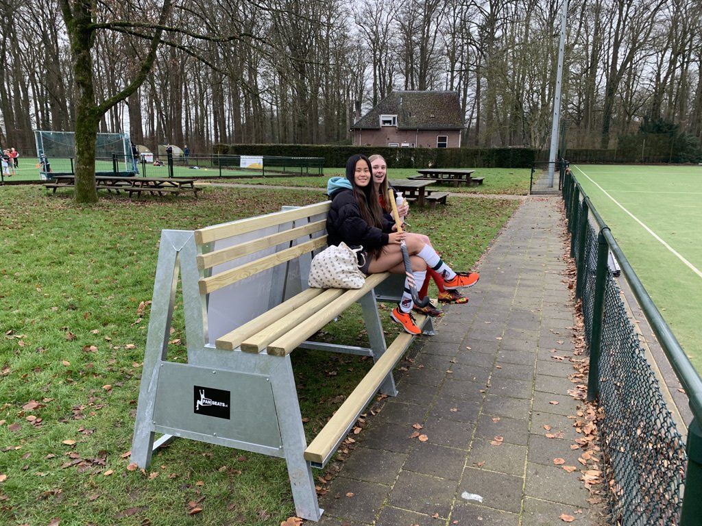 Twee mensen zitten op een Fanseats bankje naast een sportveld. De ene draagt ​​een jas, de andere hockeykleding.
