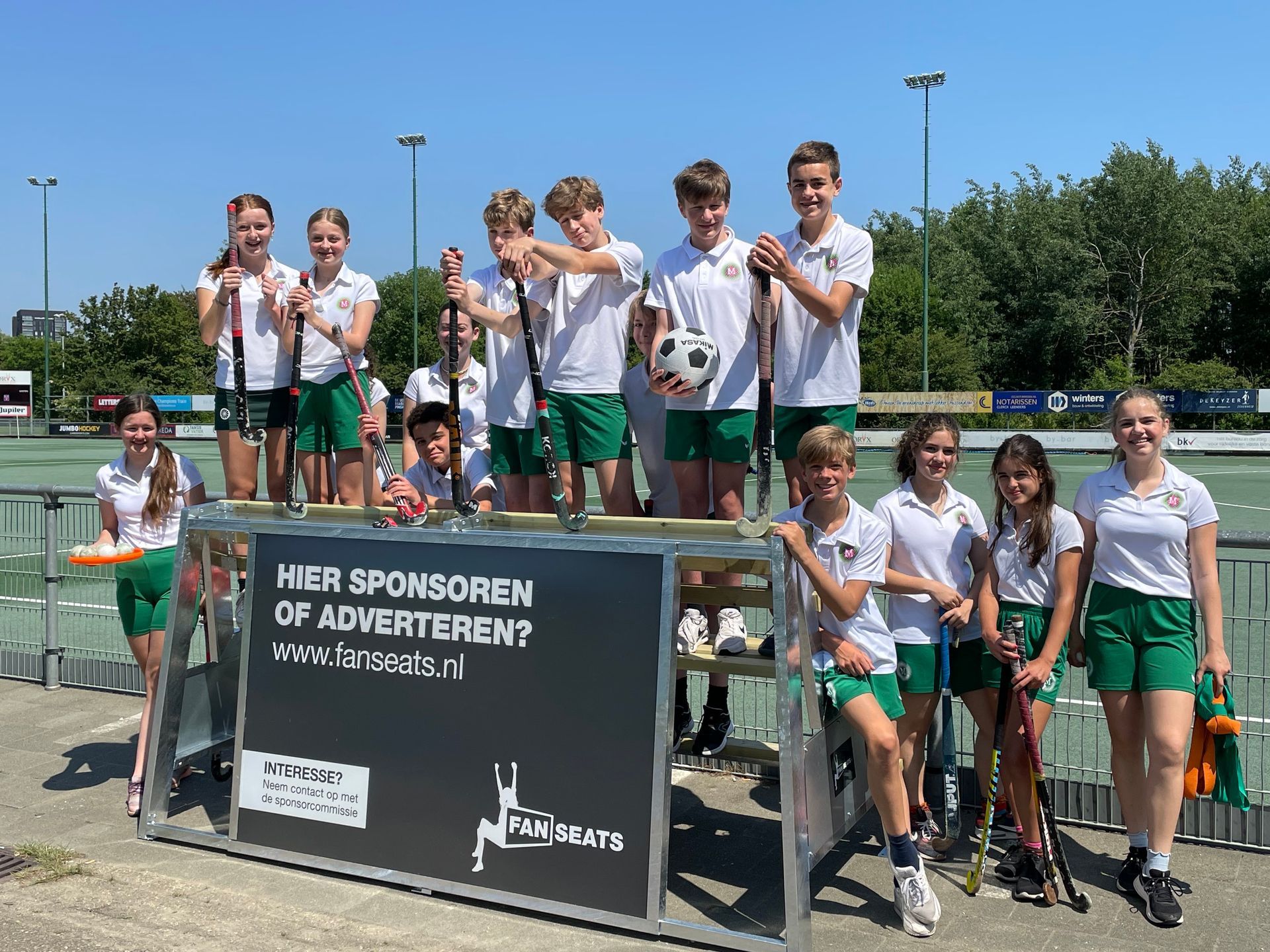 Kinderen in groene en witte uniformen poseren met hockeysticks, sommigen met een voetbal, bij een Fanseats bank.