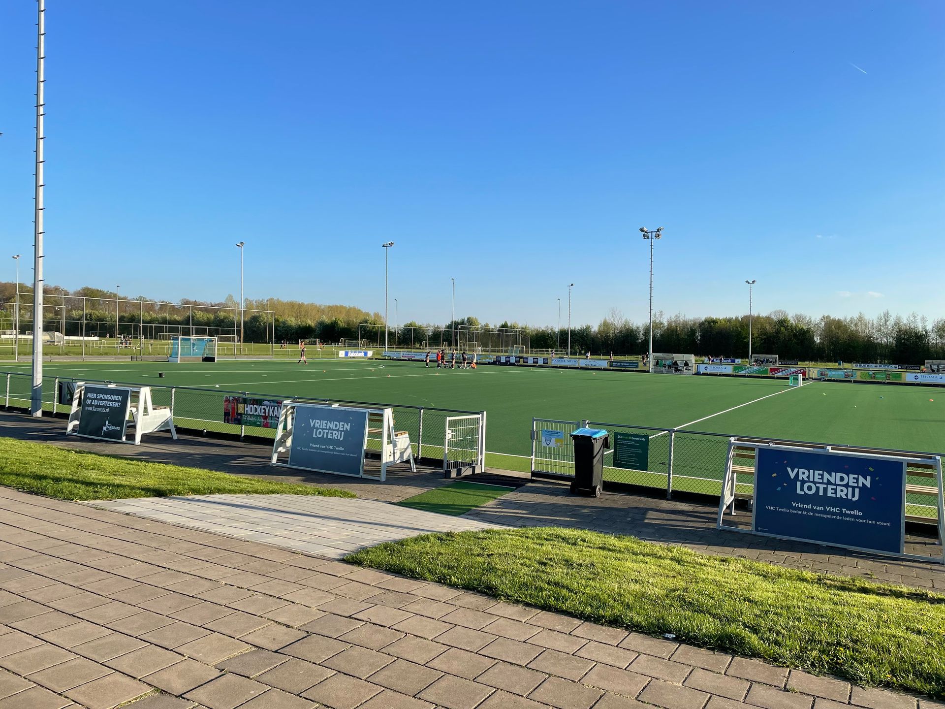Groen sportveld met banken en reclame langs het veld en een zonnige hemel.