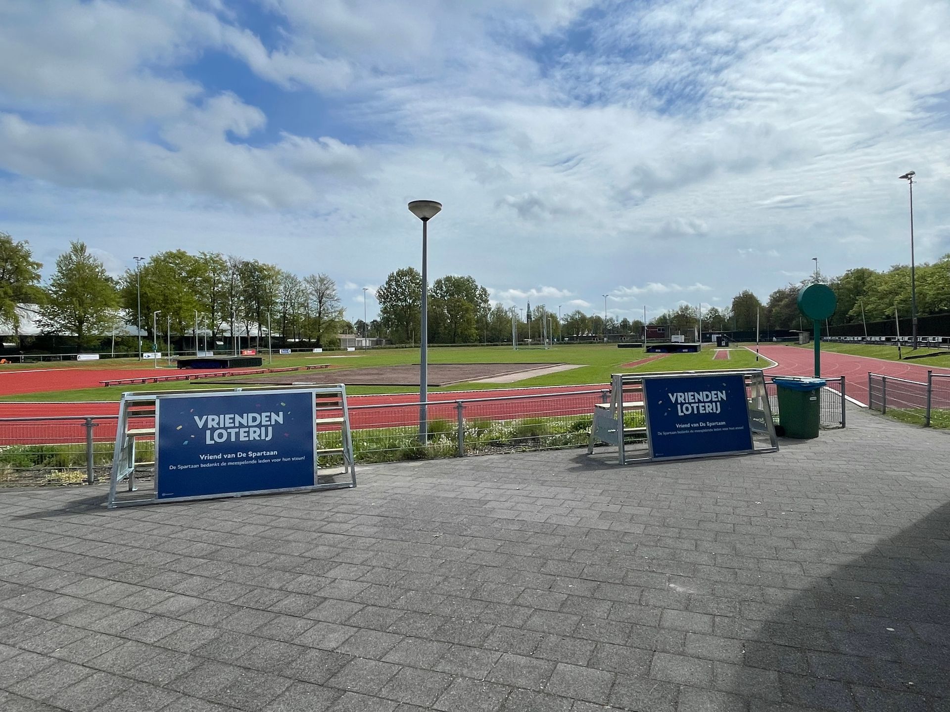 Buitensportveld met atletiekbaan, banken met reclame, onder een bewolkte hemel.