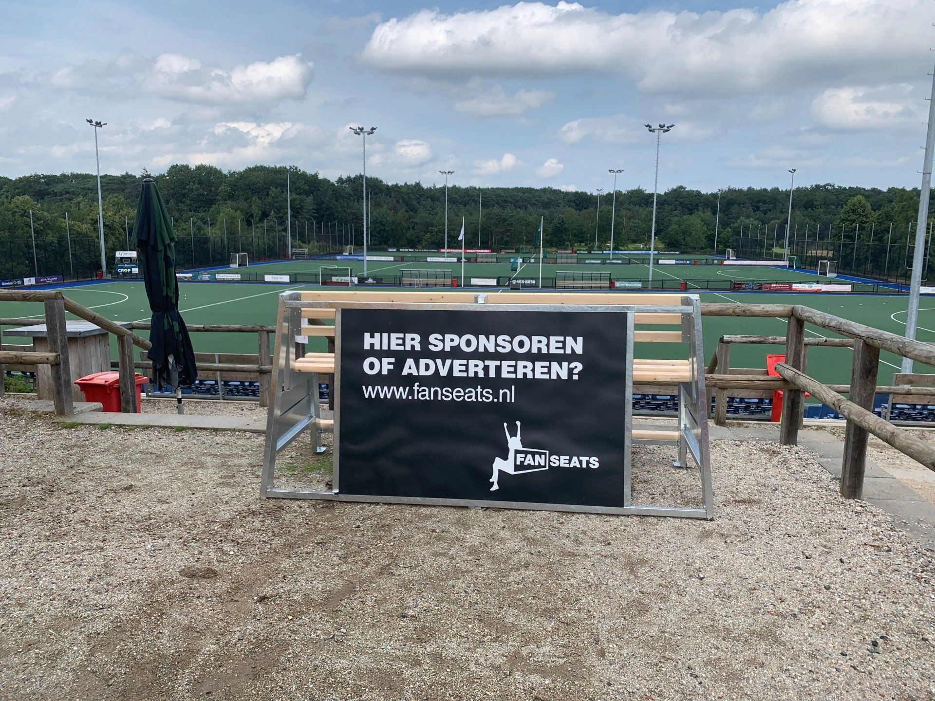 Een Fanseats bank bij hockeyveld Schaerweijde met de oproep tot sponsoring, met op de achtergrond het veld.