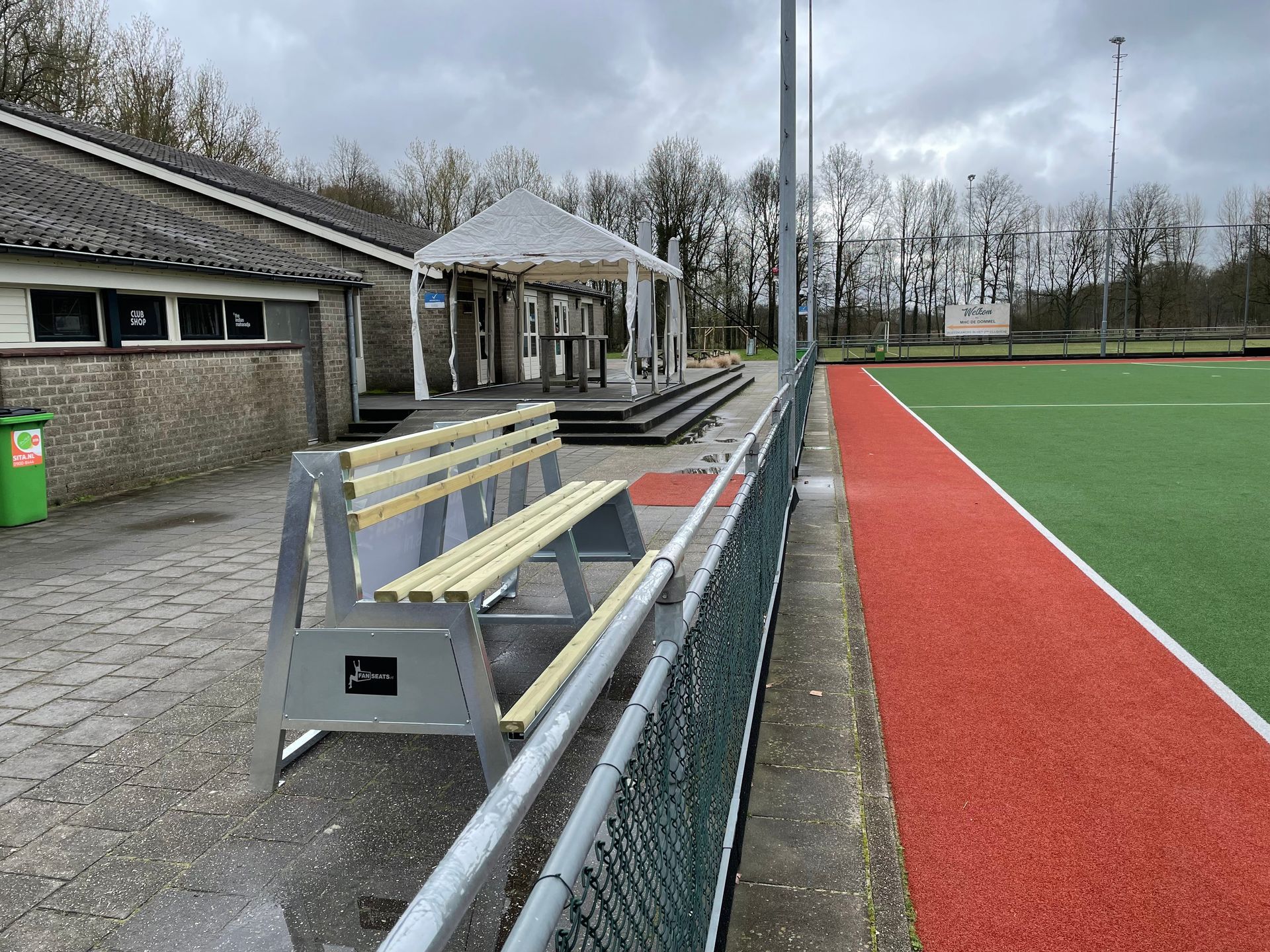 Een Fanseats bank naast een hockeyveld met een rood/groen oppervlak, vlakbij een gebouw en een afdak, op een bewolkte dag.