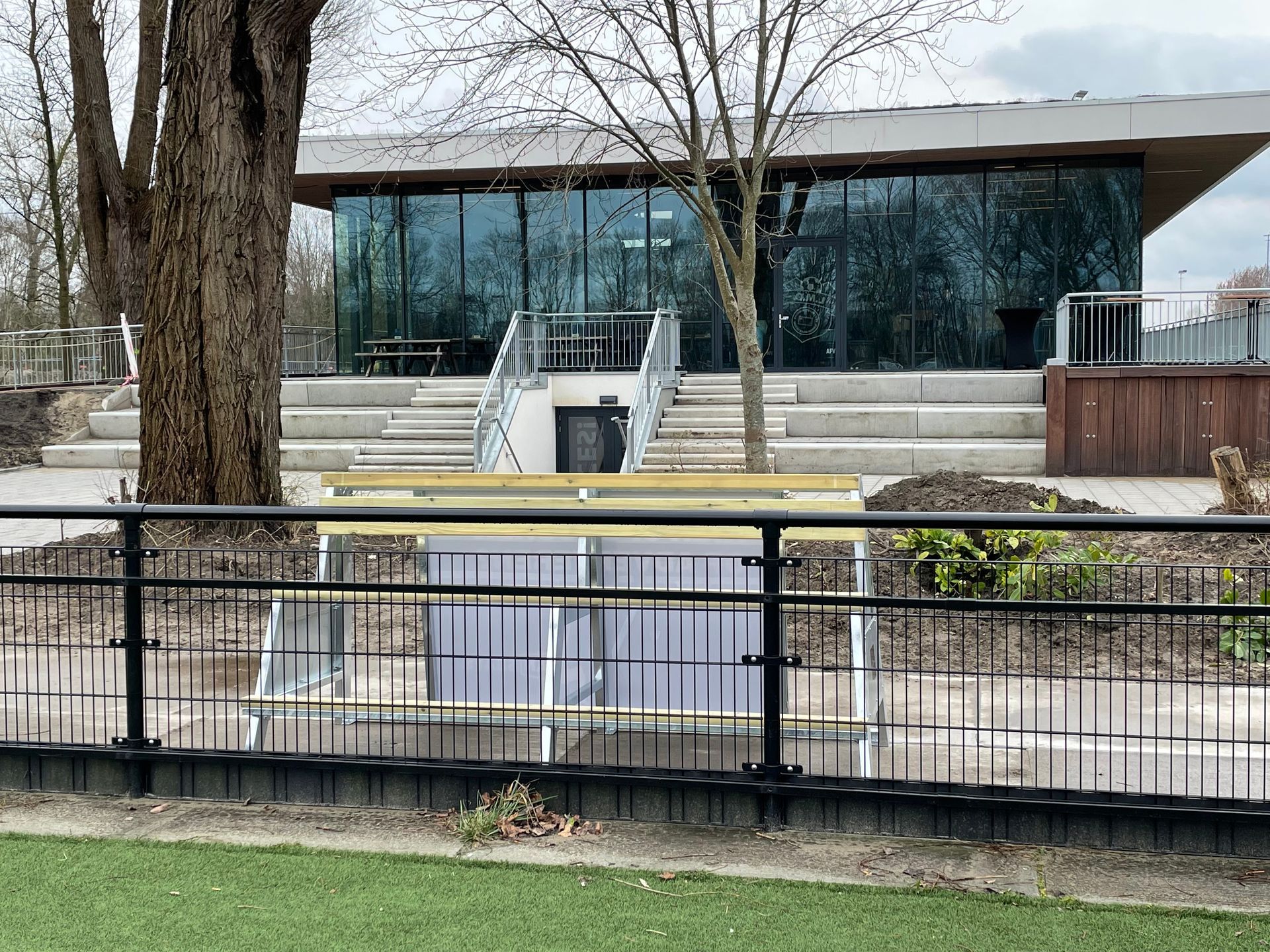 Gebouw met glazen wanden op een sportterrein, een trap naar boven en een zwart hek ervoor.