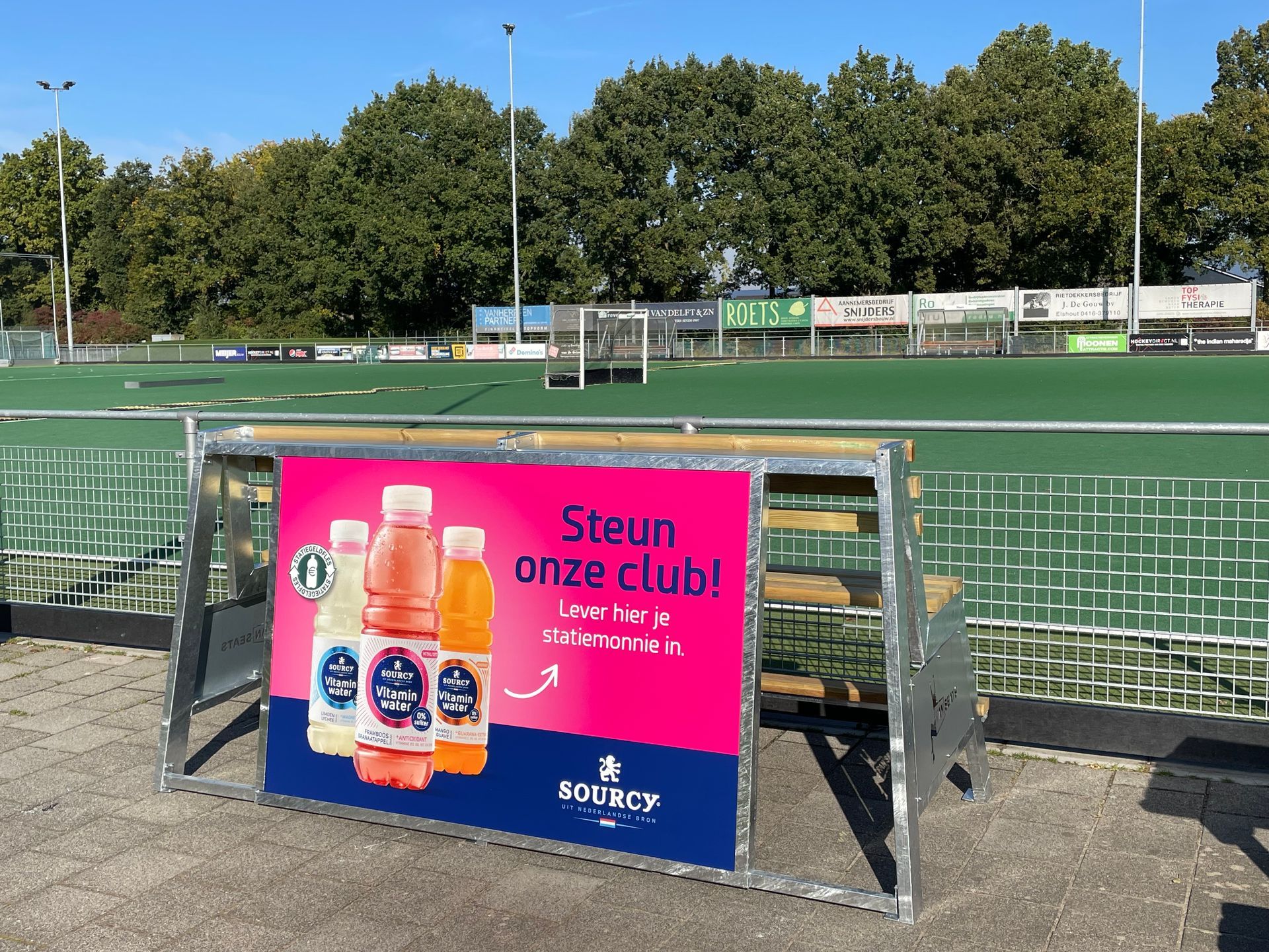 Een Fanseats bank met reclame van Sourcy Vitaminwater ter promotie van statiegeld staat voor een hockeyveld.
