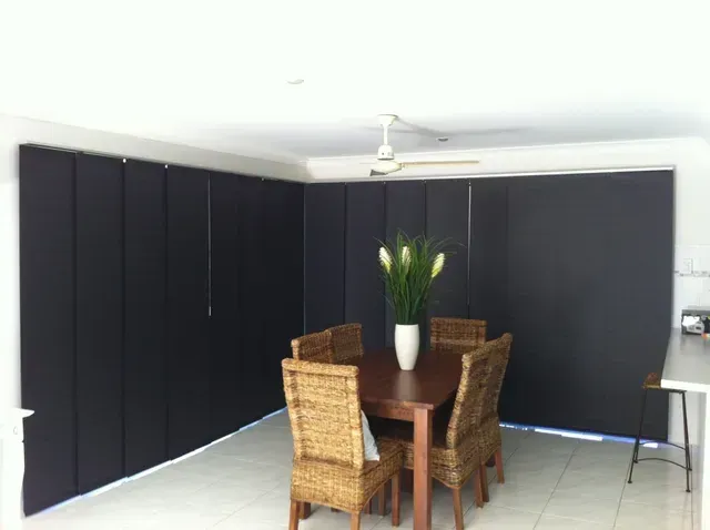 Dark blinds cover the windows of a dining area with a table, chairs, and a flower vase.