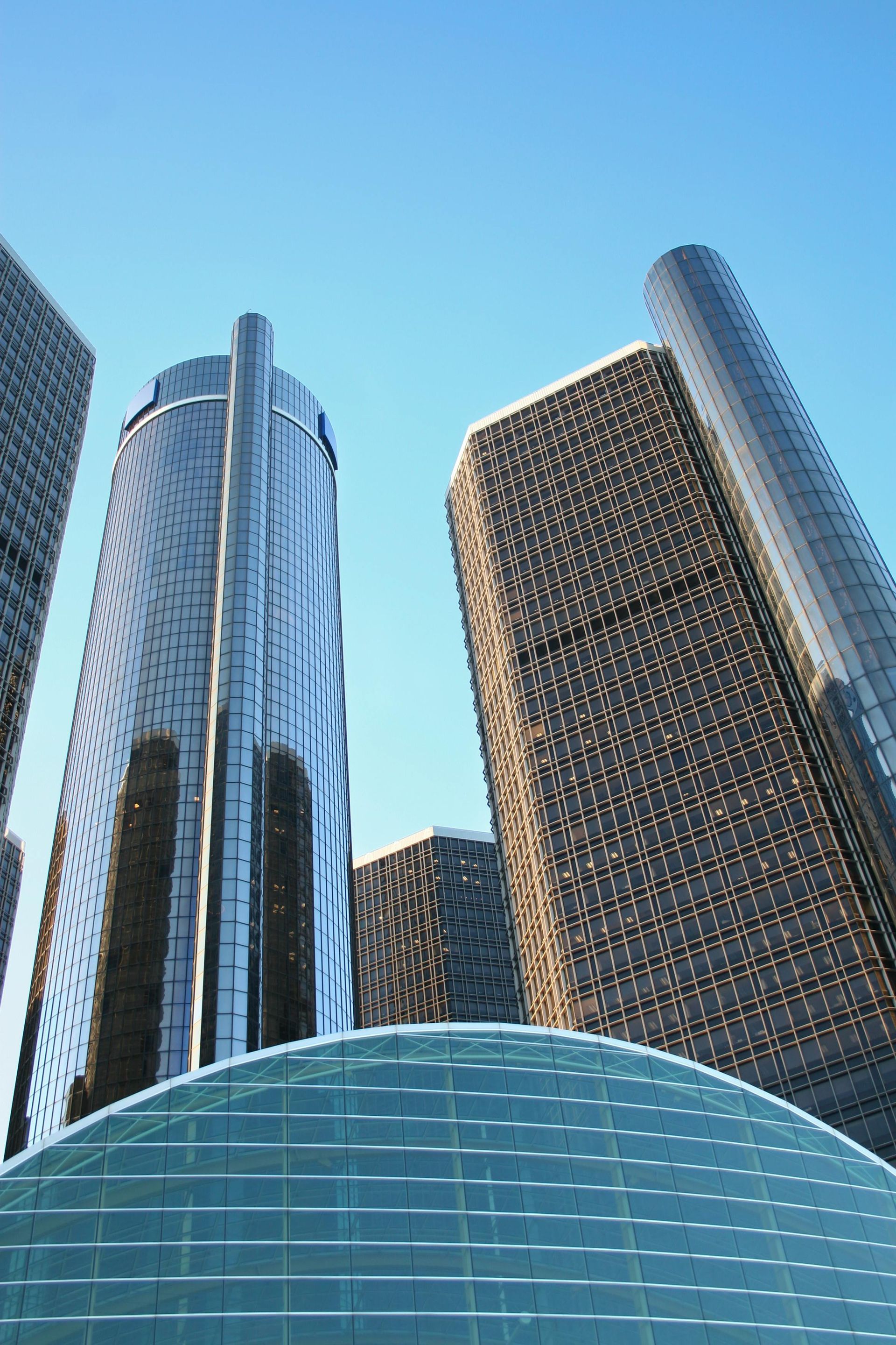The Renaissance Center skyscrapers rise against a clear blue sky, viewed from a low angle above a curved glass roof.