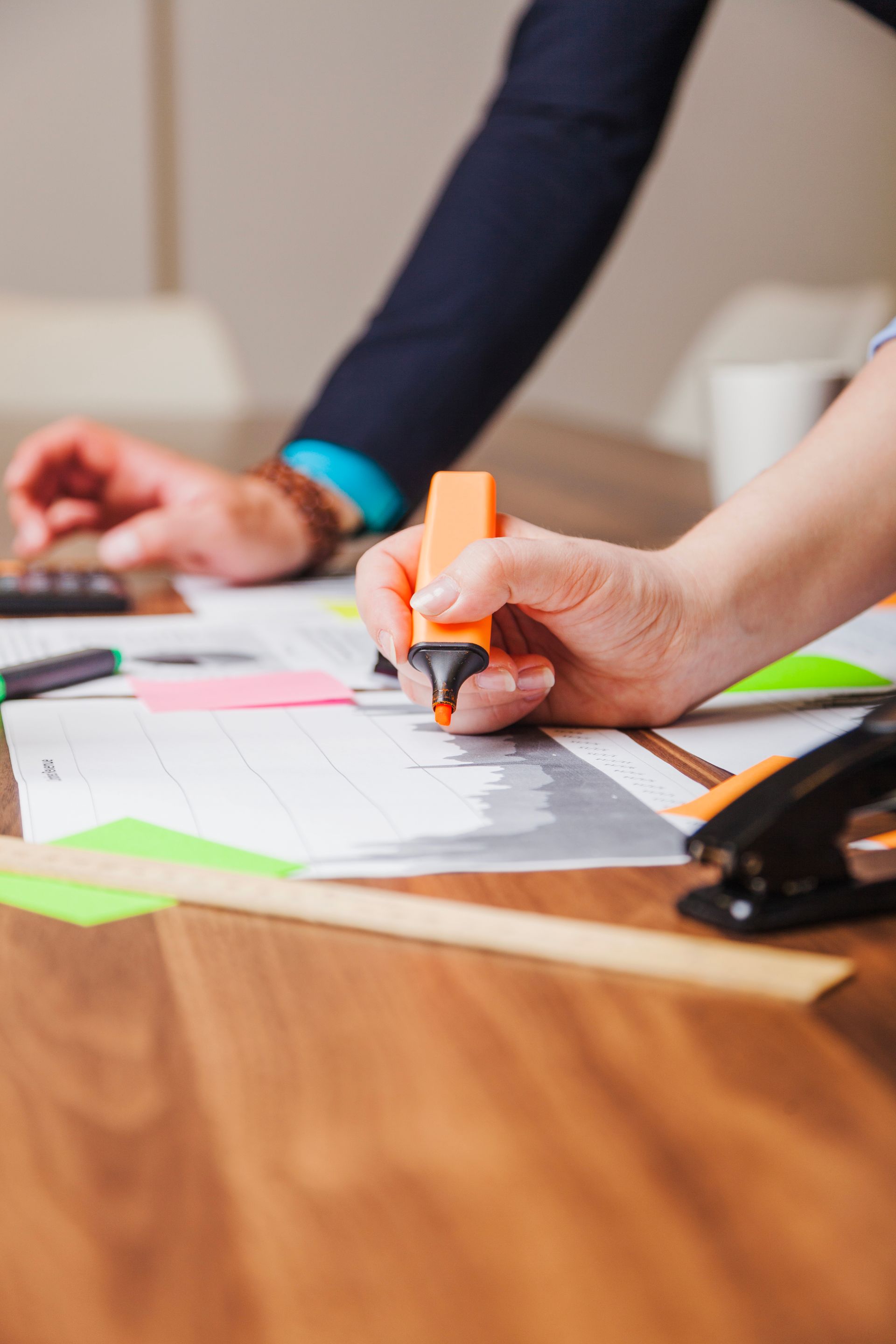 A person holding an orange highlighter over papers on a wooden desk with a stapler, calculator, and ruler nearby.