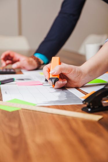 A person holding an orange highlighter over papers on a wooden desk with a stapler, calculator, and ruler nearby.