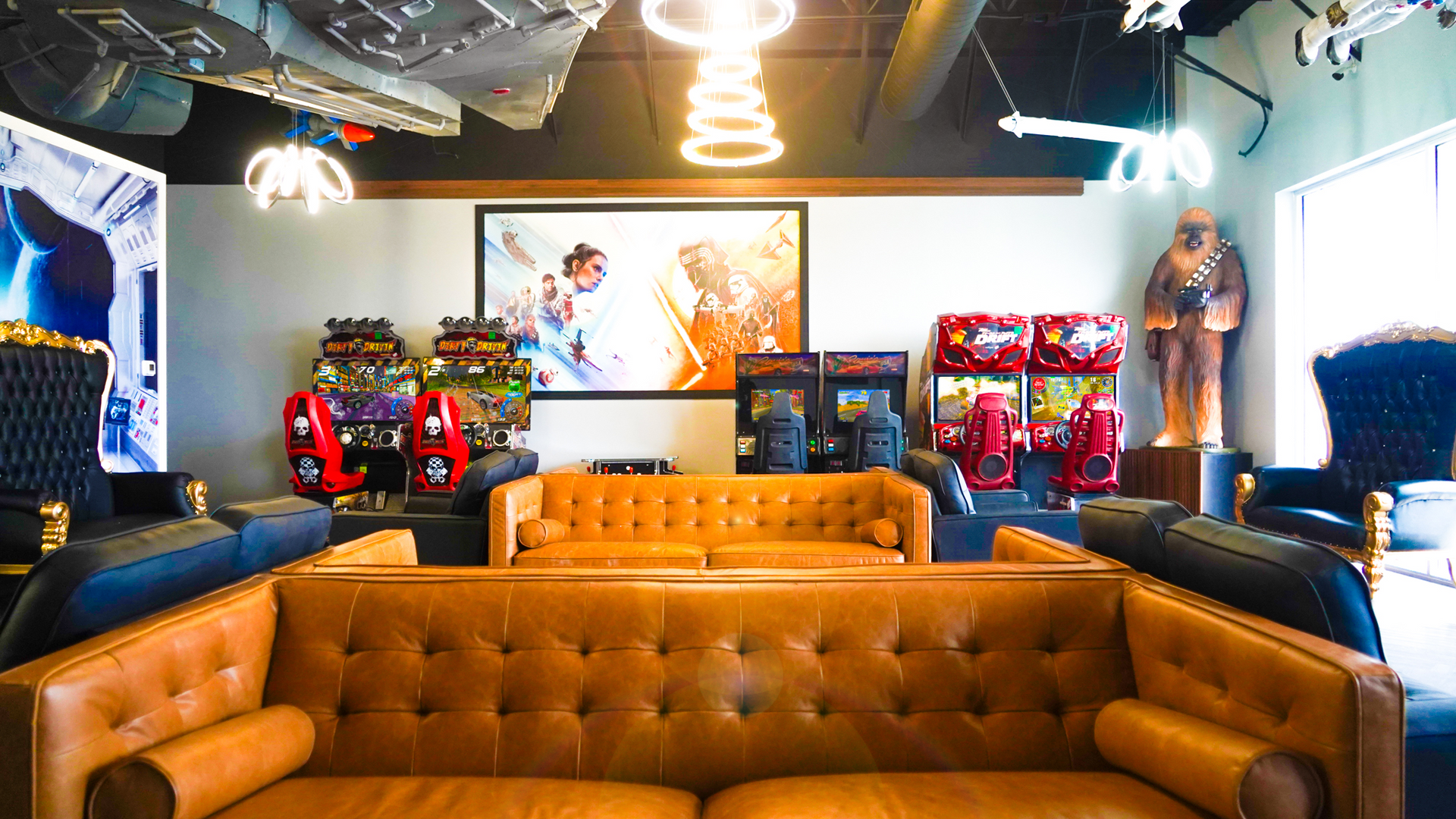 A lounge with tan leather sofas, arcade games, and Star Wars decor, including a Chewbacca statue and neon lighting.