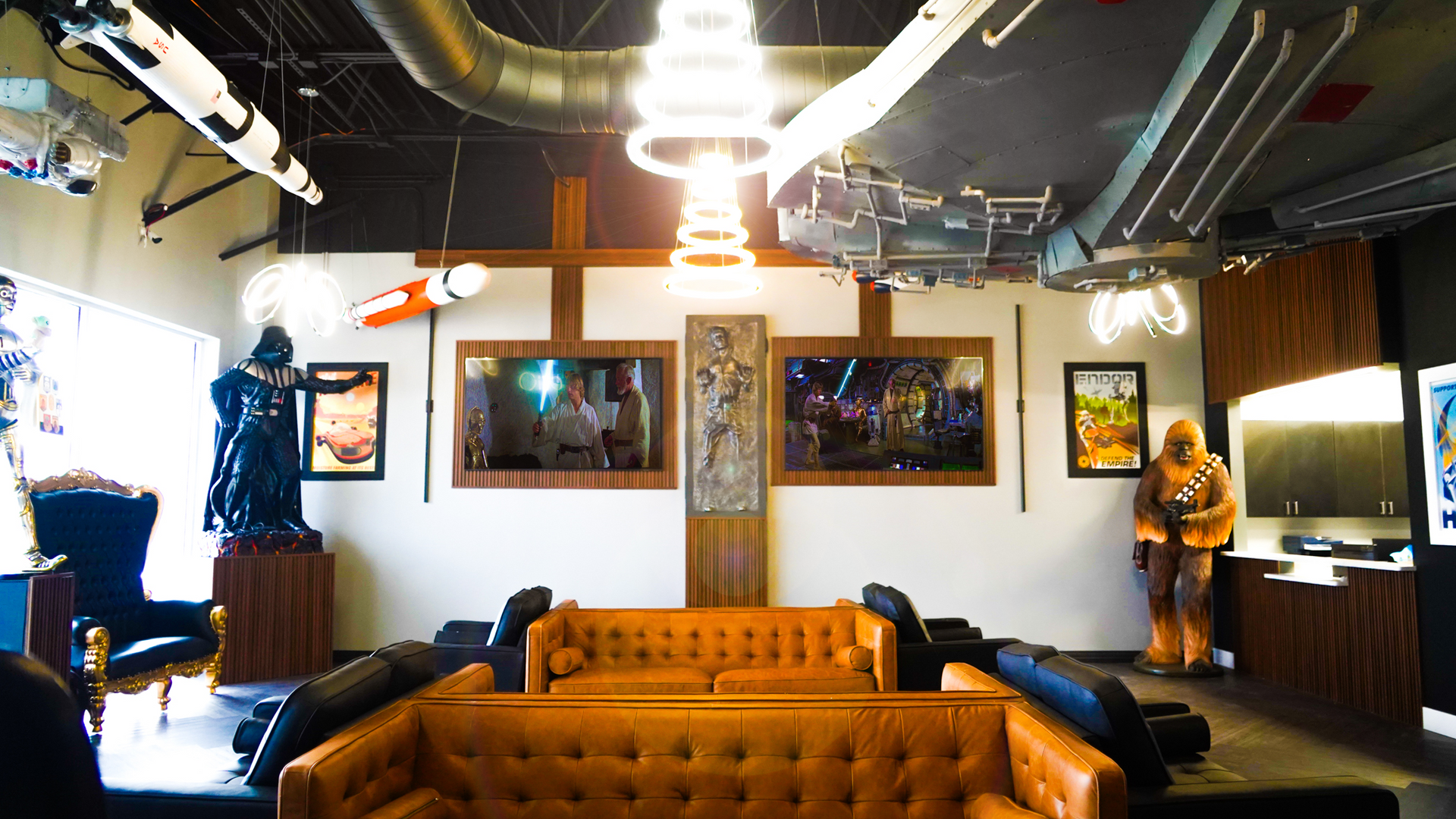 A Star Wars-themed room featuring a Darth Vader statue, a Chewbacca figure, brown leather sofas, and movie posters.