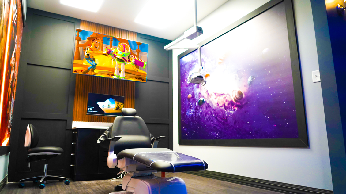 A pediatric dental office with a treatment chair, a screen showing a cartoon, and a large space-themed mural on the wall.