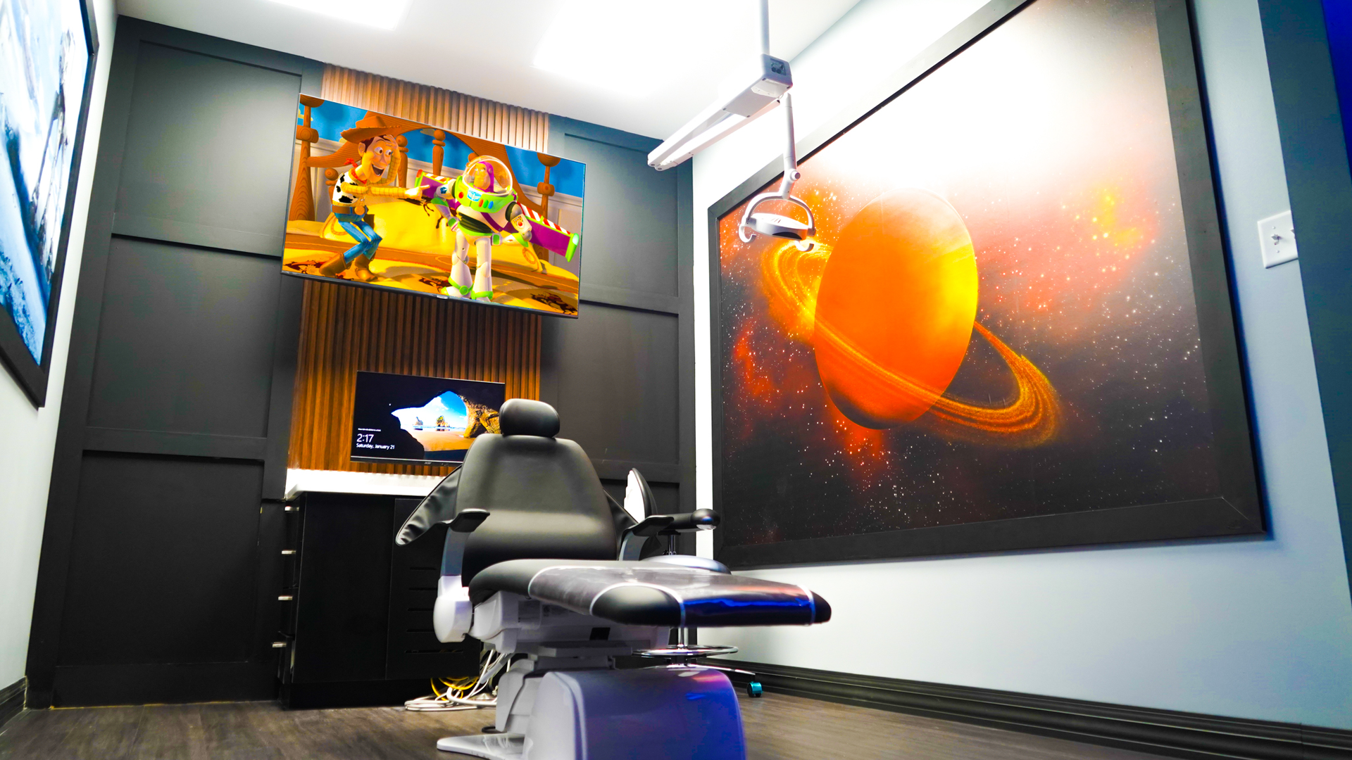 A dental exam room with a central chair, a flat-screen TV displaying Toy Story, and a large mural of Saturn on the wall.