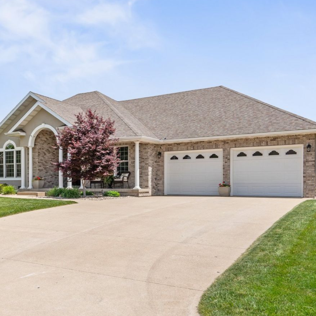 Two-car garage attached to a brick house with a front porch and a curving driveway on a sunny day.