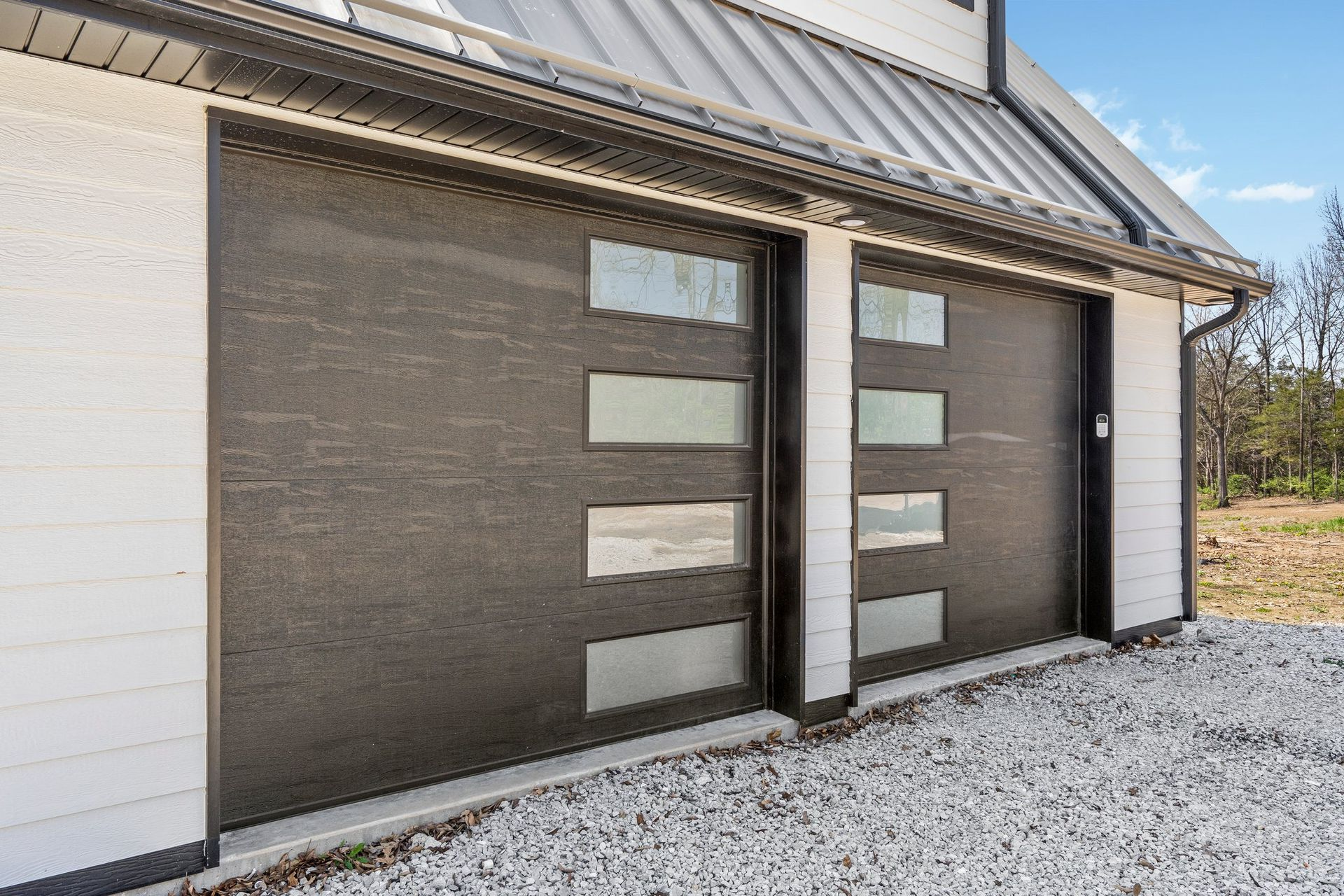 Two dark garage doors with rectangular glass windows, set in a white building with gravel.