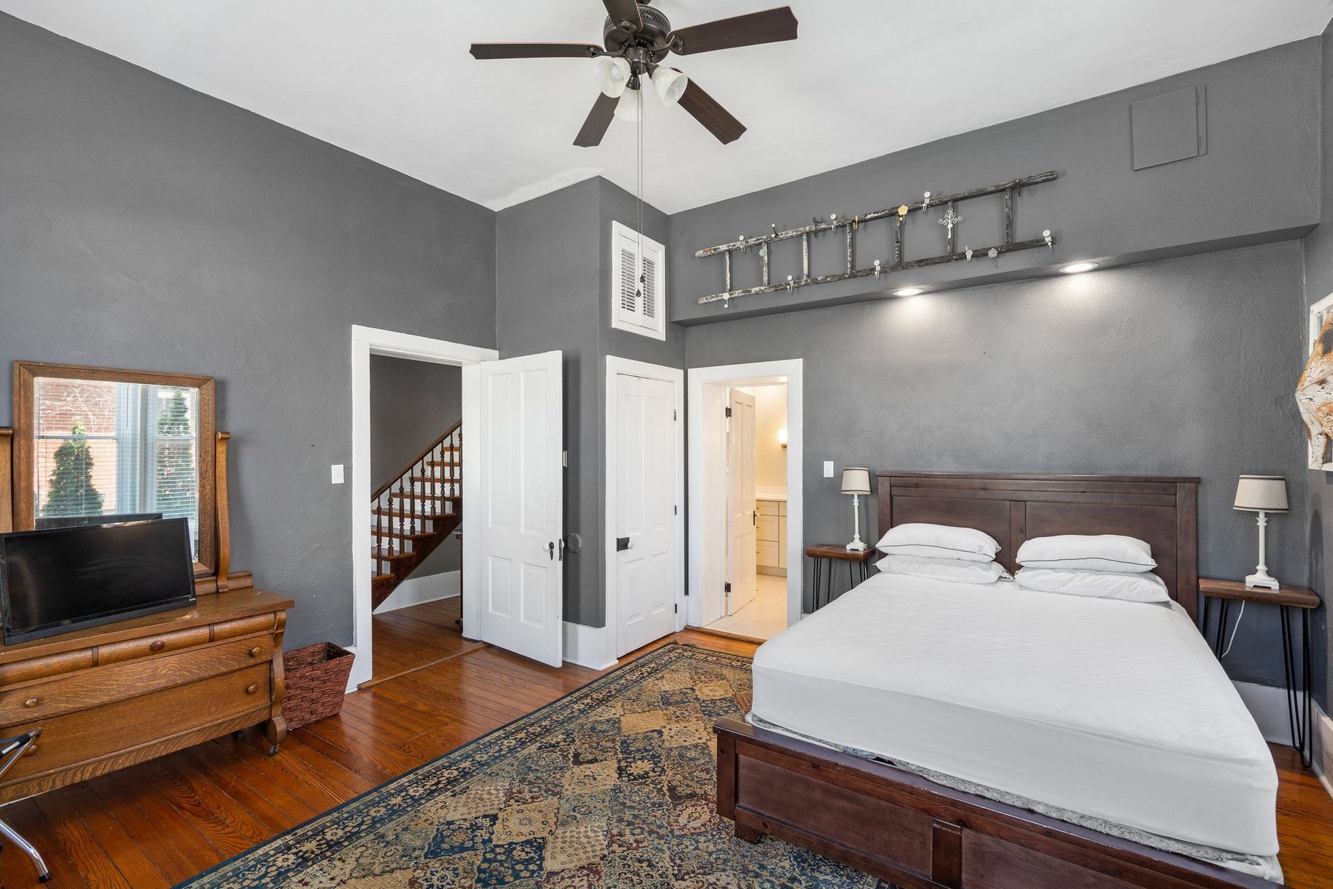 Bedroom with gray walls, wooden bed frame, rug, and ladder decor. Doorways lead to stairs and bathroom.