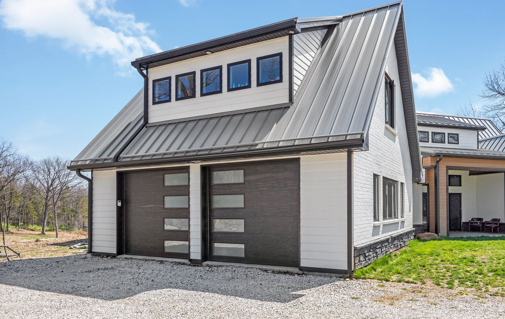 Modern white house with gray metal roof and garage doors, windows, and gravel driveway.