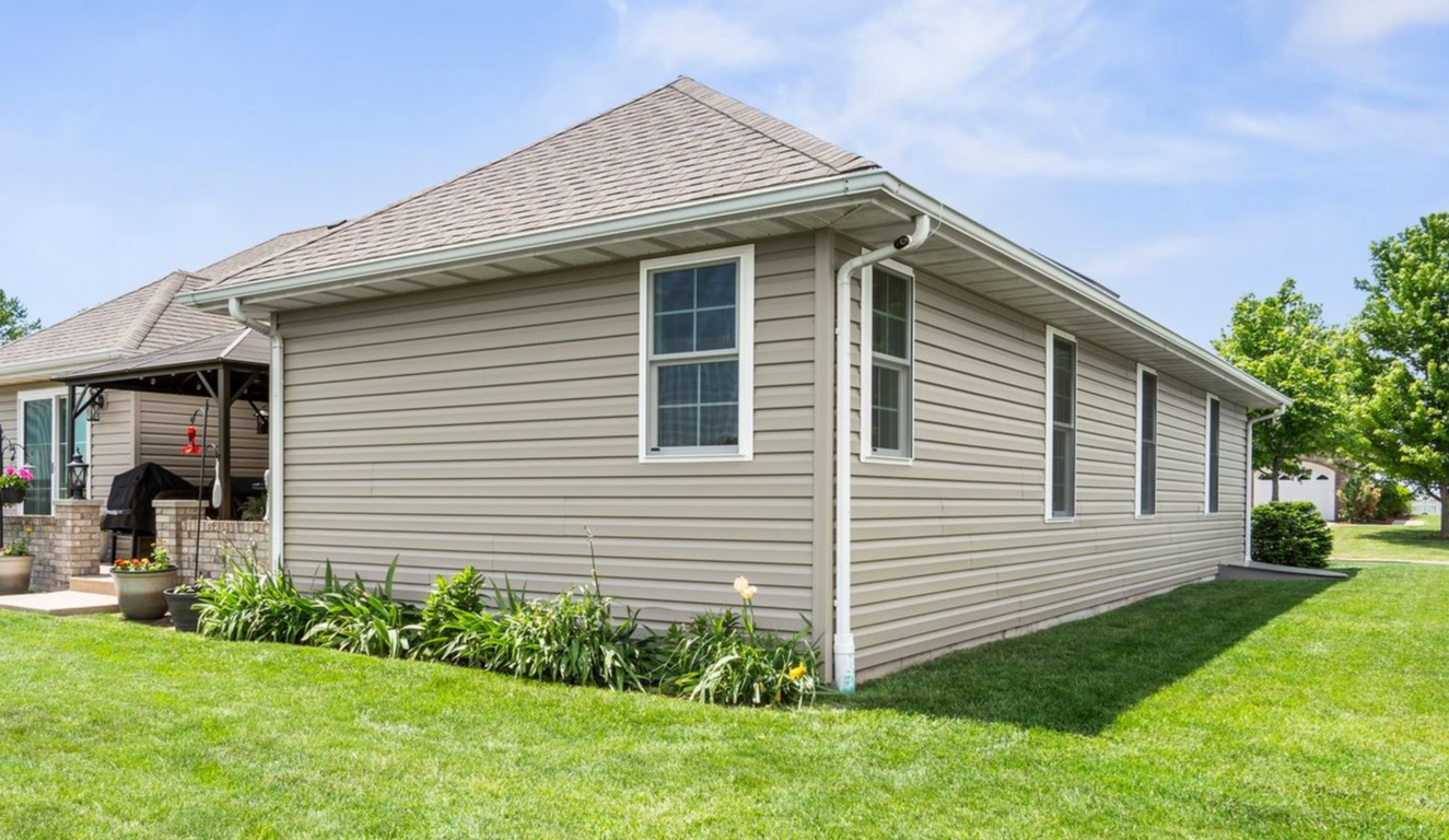 Beige house with a brown roof and white trim, surrounded by green grass and trees.