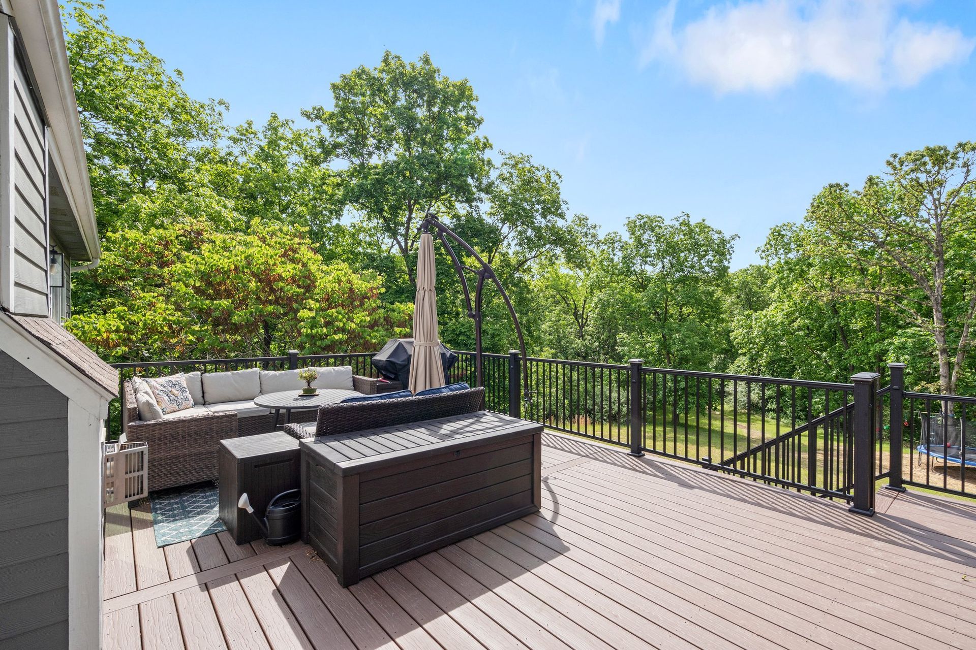Backyard deck with wicker furniture, grill, and surrounding trees under a blue sky.
