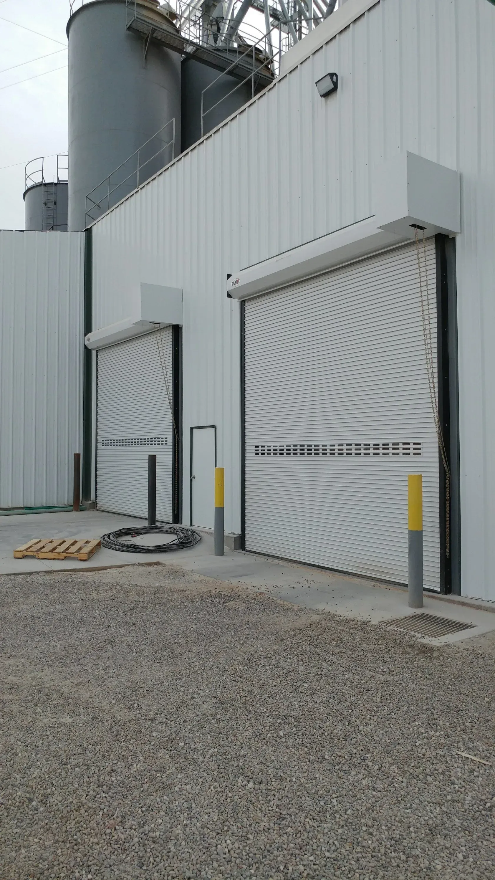 White industrial building with two closed roll-up doors, gravel ground, and grain silos in the background.