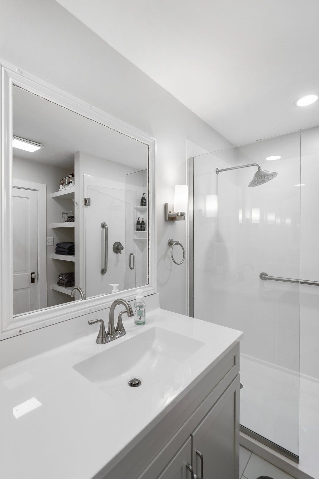 White bathroom with vanity, shower, and accessible grab bars. Mirror reflects shelving and a door.