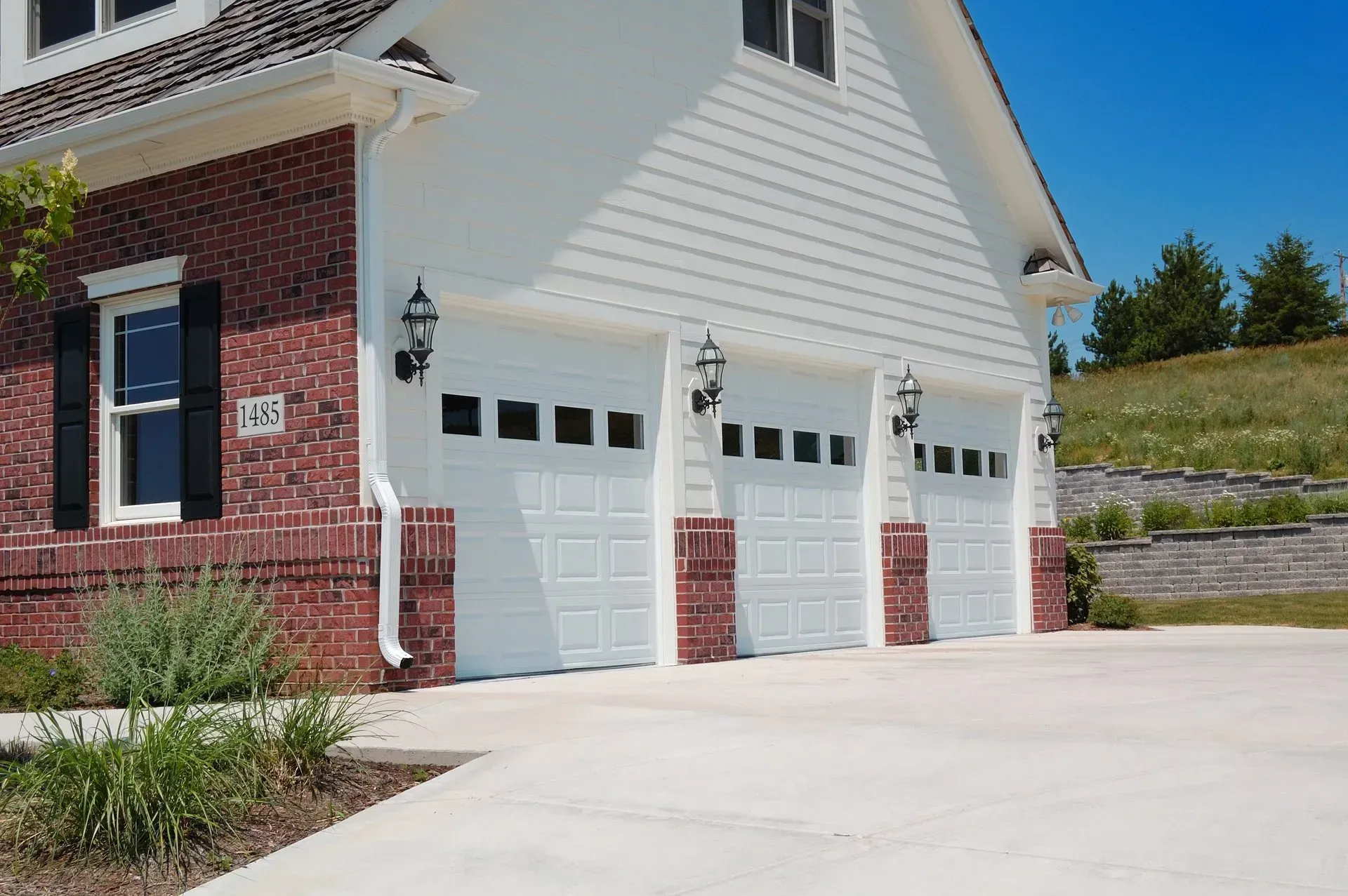 Three-car garage with white doors, red brick facade, and white siding. Clear blue sky.