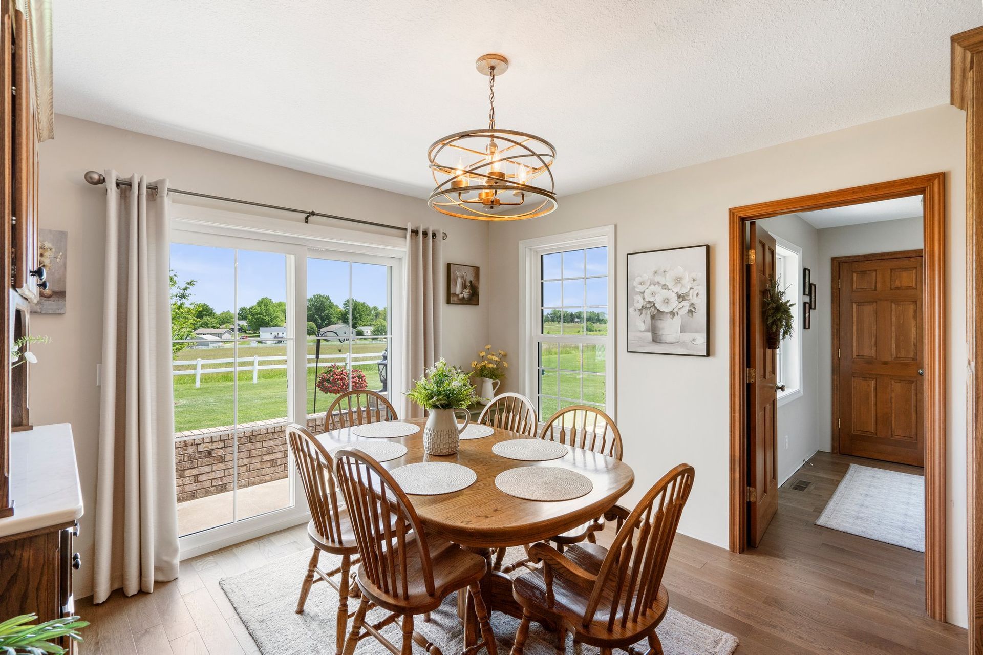 Dining room with wooden table, chairs, and chandelier, overlooking a grassy field.