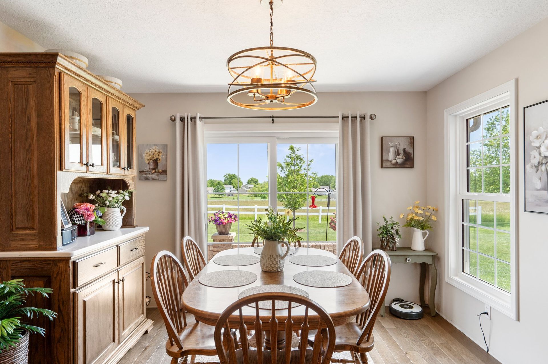 Dining room with wooden table, chairs, hutch, and large windows overlooking a grassy field.