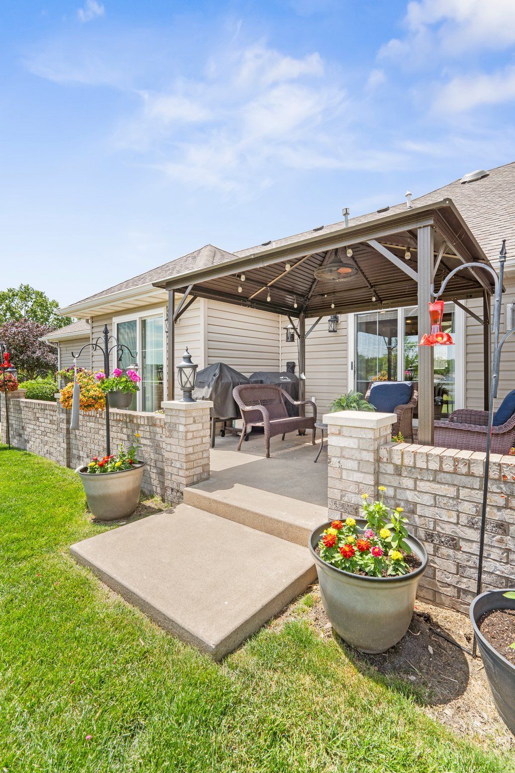 Patio with brick pillars, seating, and a pergola, surrounded by grass and flower pots.