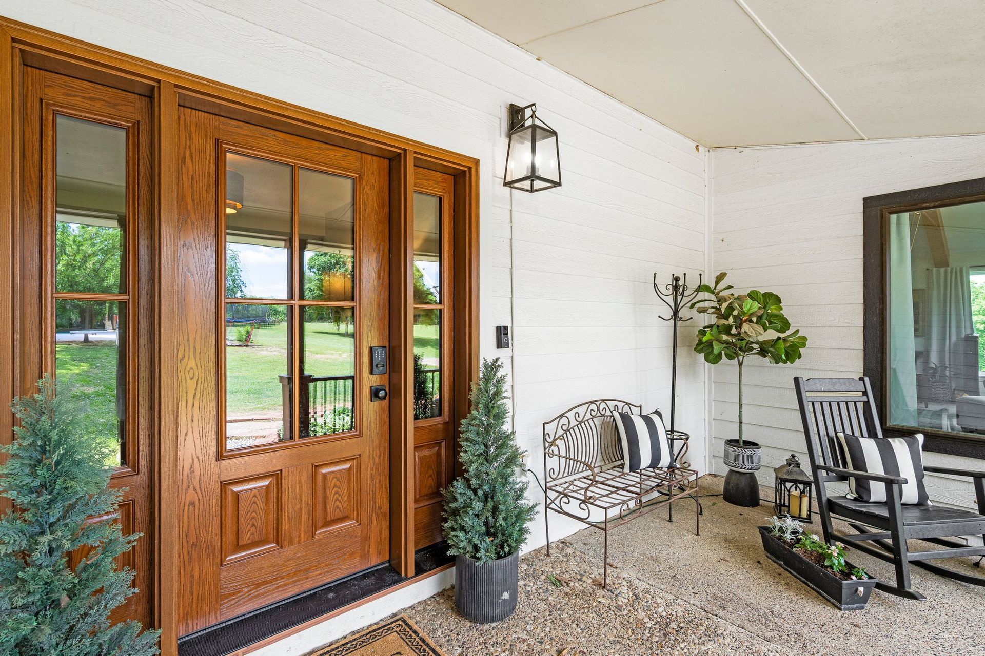 Wooden front door on a porch with seating, small trees, and a view of a yard.