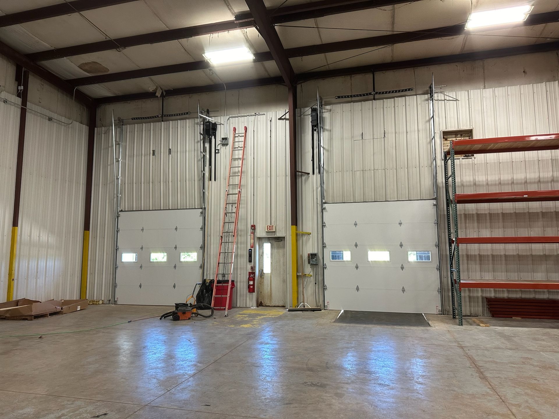 Large industrial warehouse interior with tall roll-up doors, red ladder, and shelving on the right.