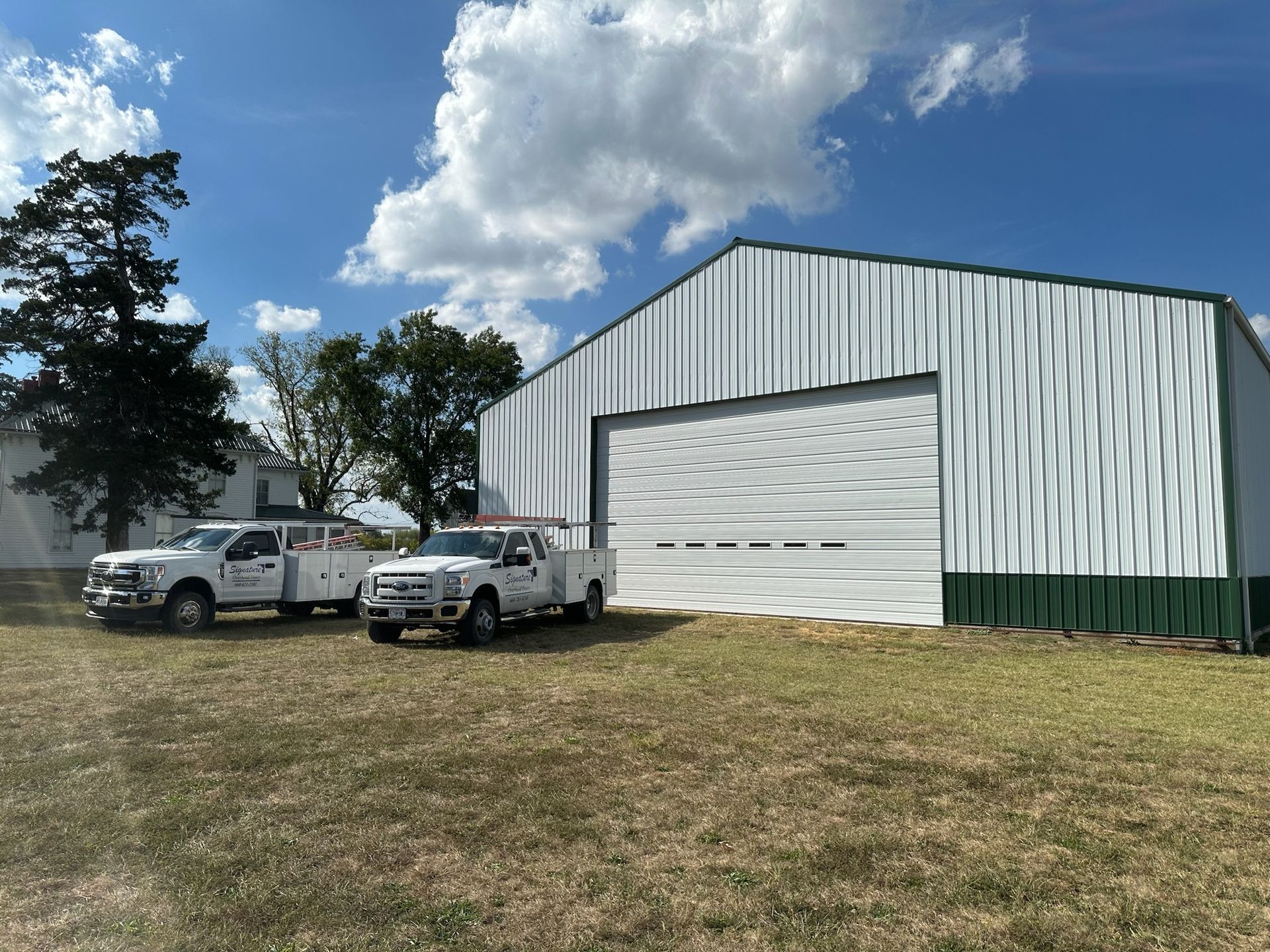 Two white trucks parked near a white metal building with a large garage door. Blue sky with clouds.