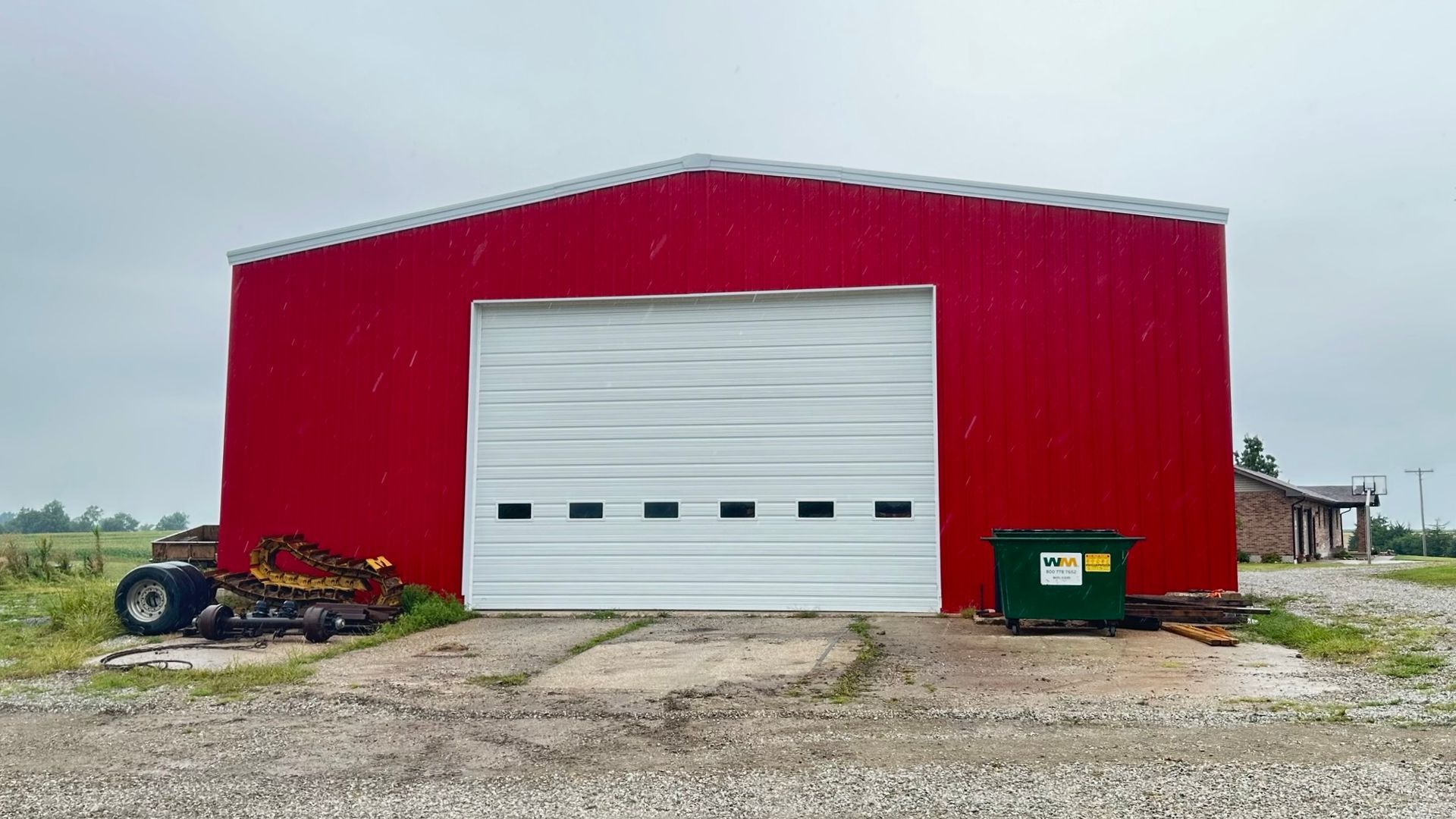 Red metal barn with large white roll-up door on a gravel lot, with a green dumpster nearby.