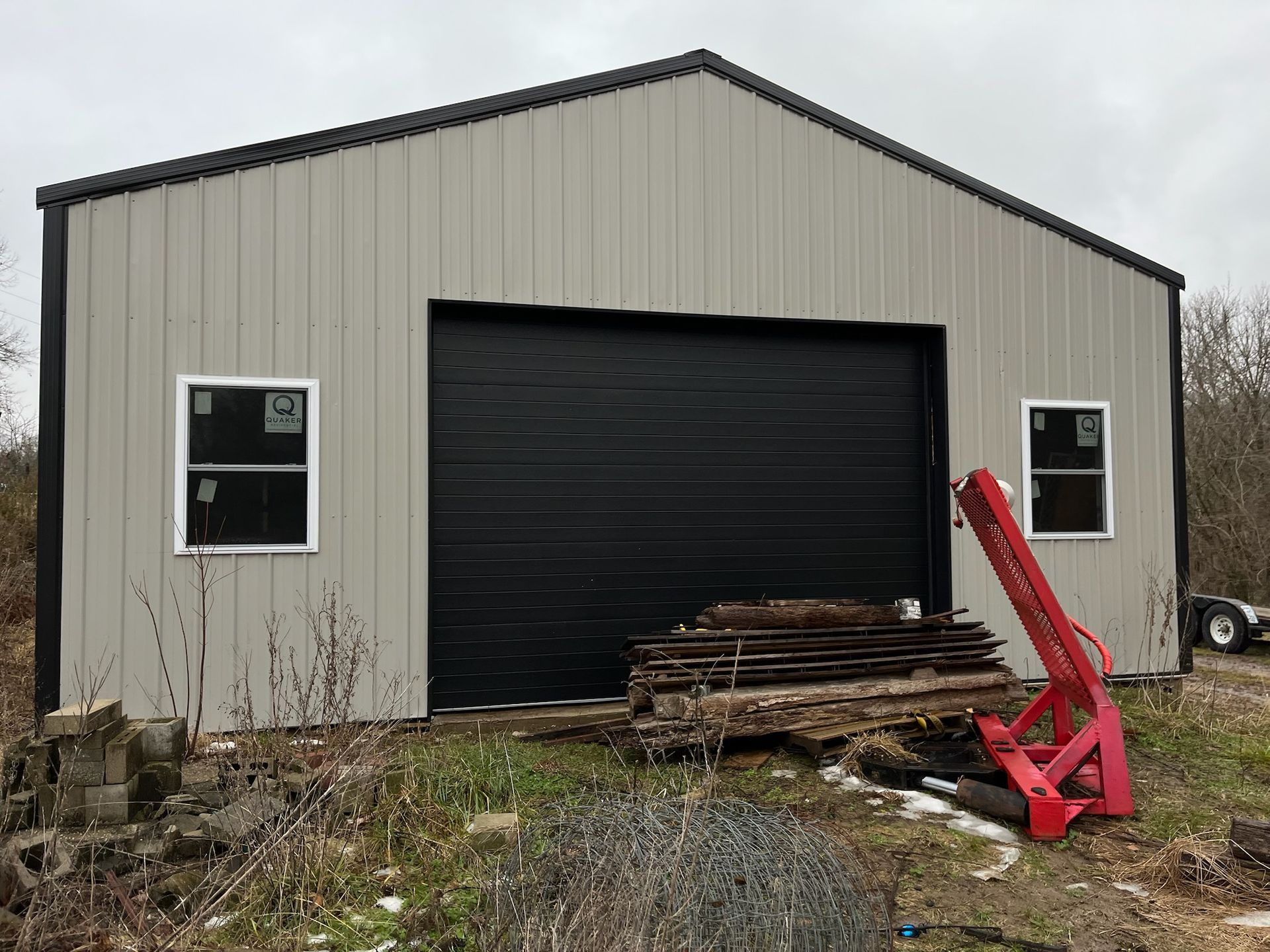 Beige metal garage with black roll-up door, two small windows, and red equipment leaning by the entrance.