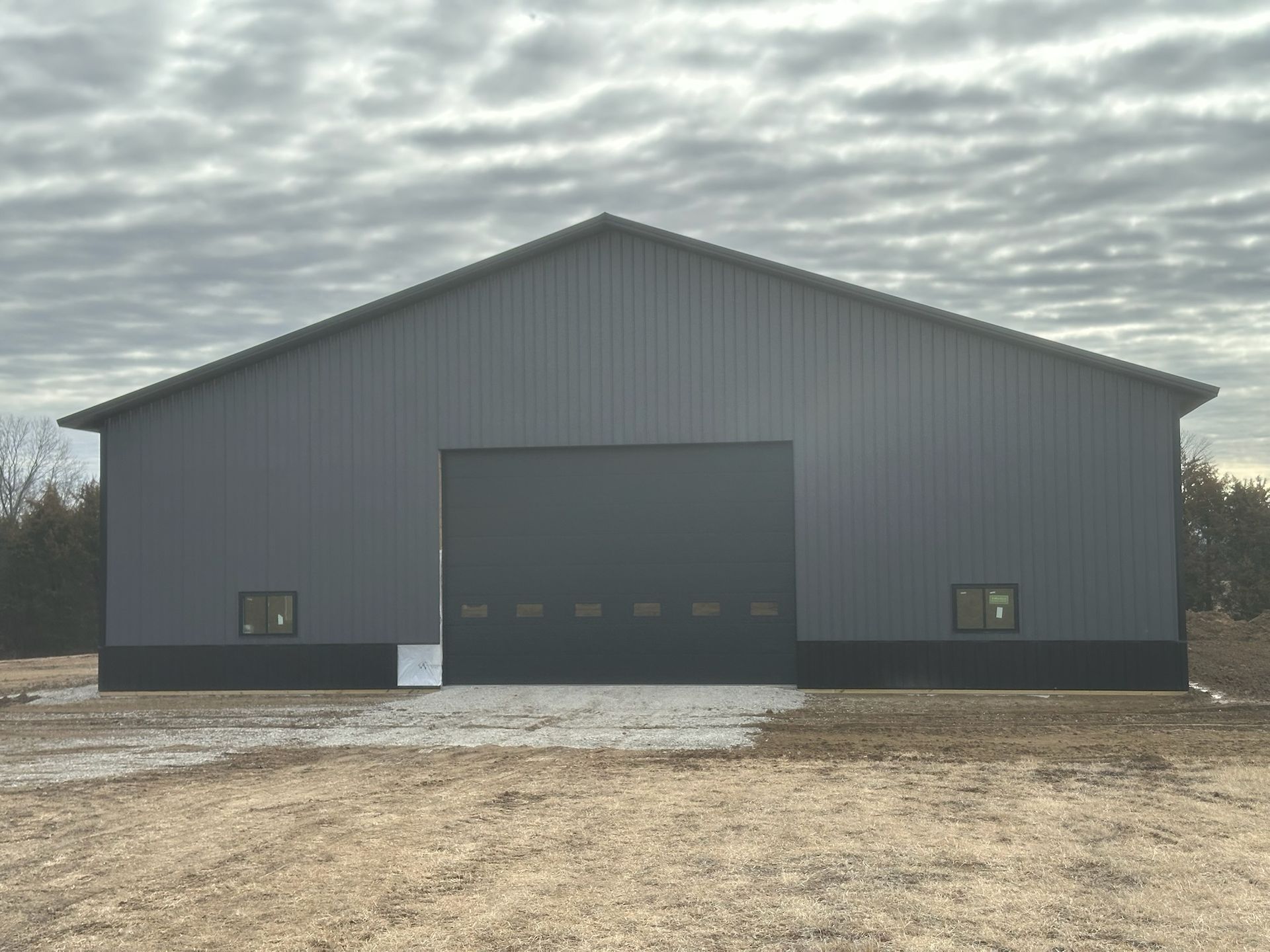 Large gray metal barn with a dark garage door in a muddy open field under cloudy sky