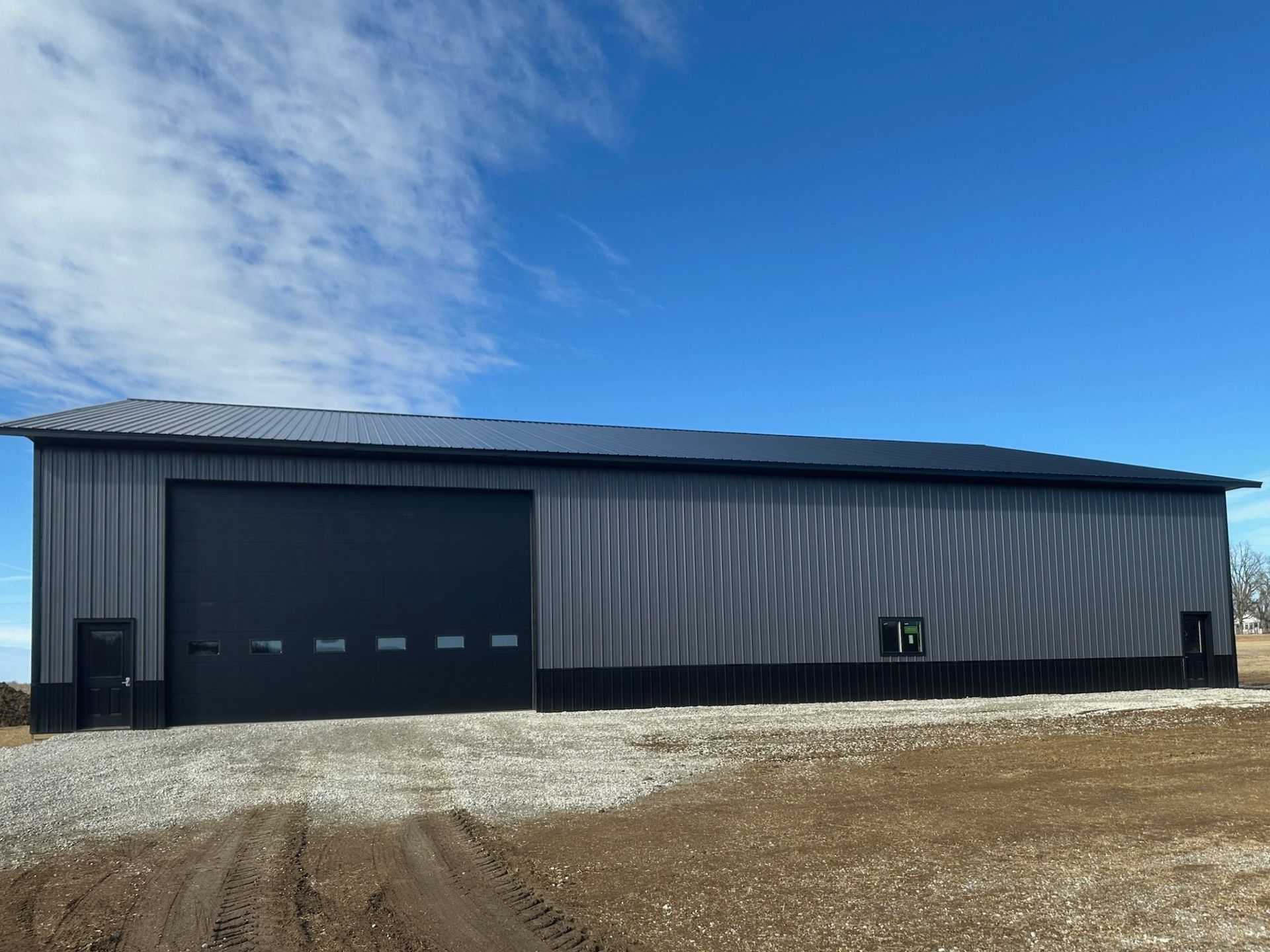 Large gray metal warehouse with a black garage door under a blue sky, on a gravel lot.