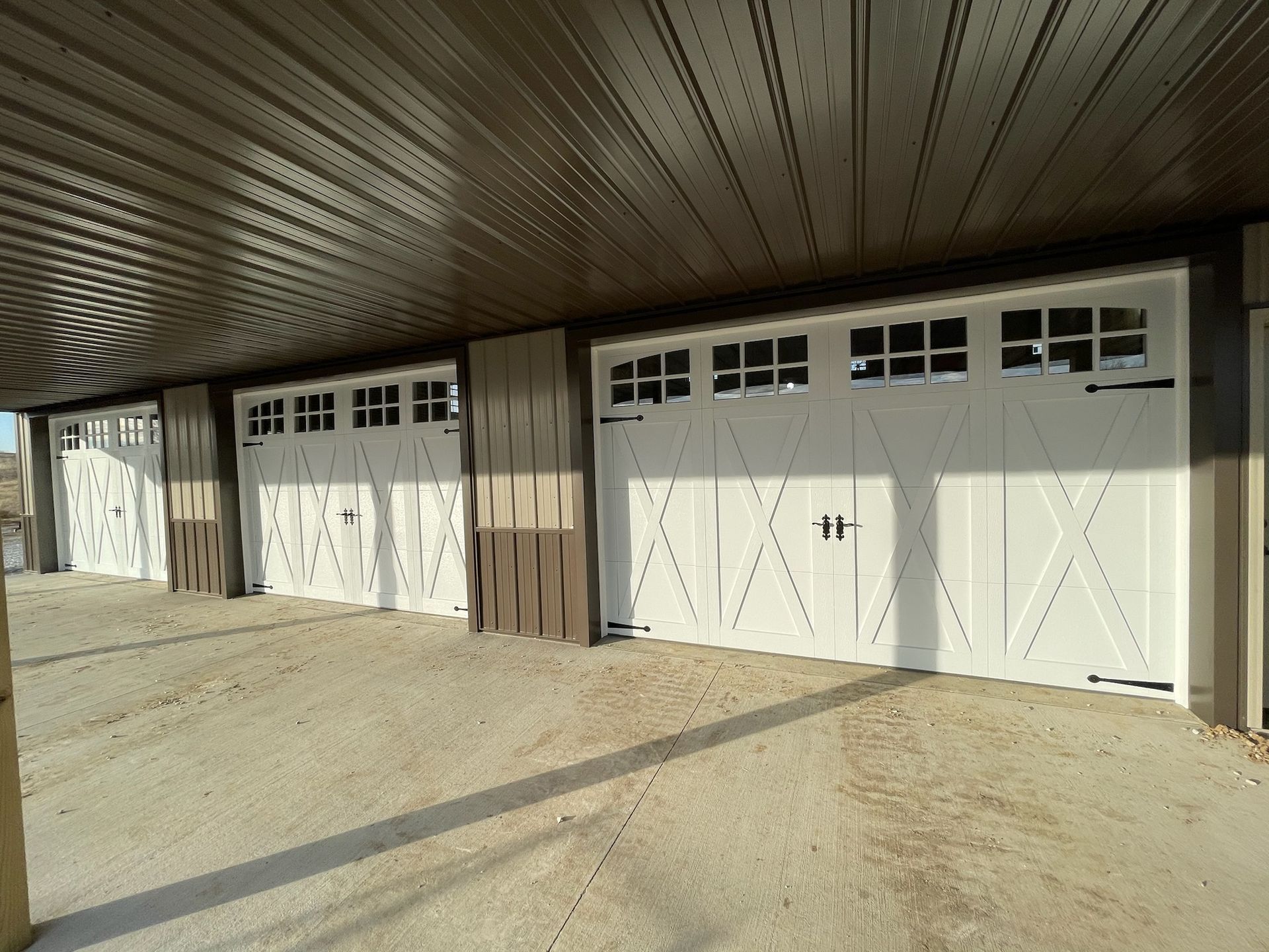 Row of white garage doors under a brown metal roof beside a dirt driveway