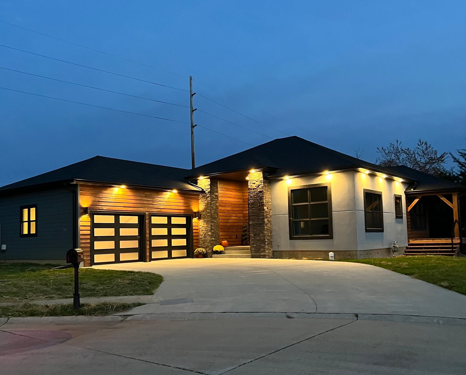 Modern house at dusk, lit up exterior, concrete driveway, and garage door.