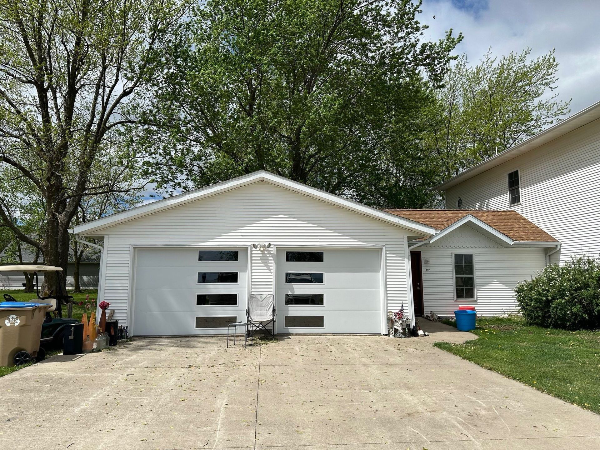 White two-car garage with modern glass-panel doors. A driveway, trees, and partial house visible.