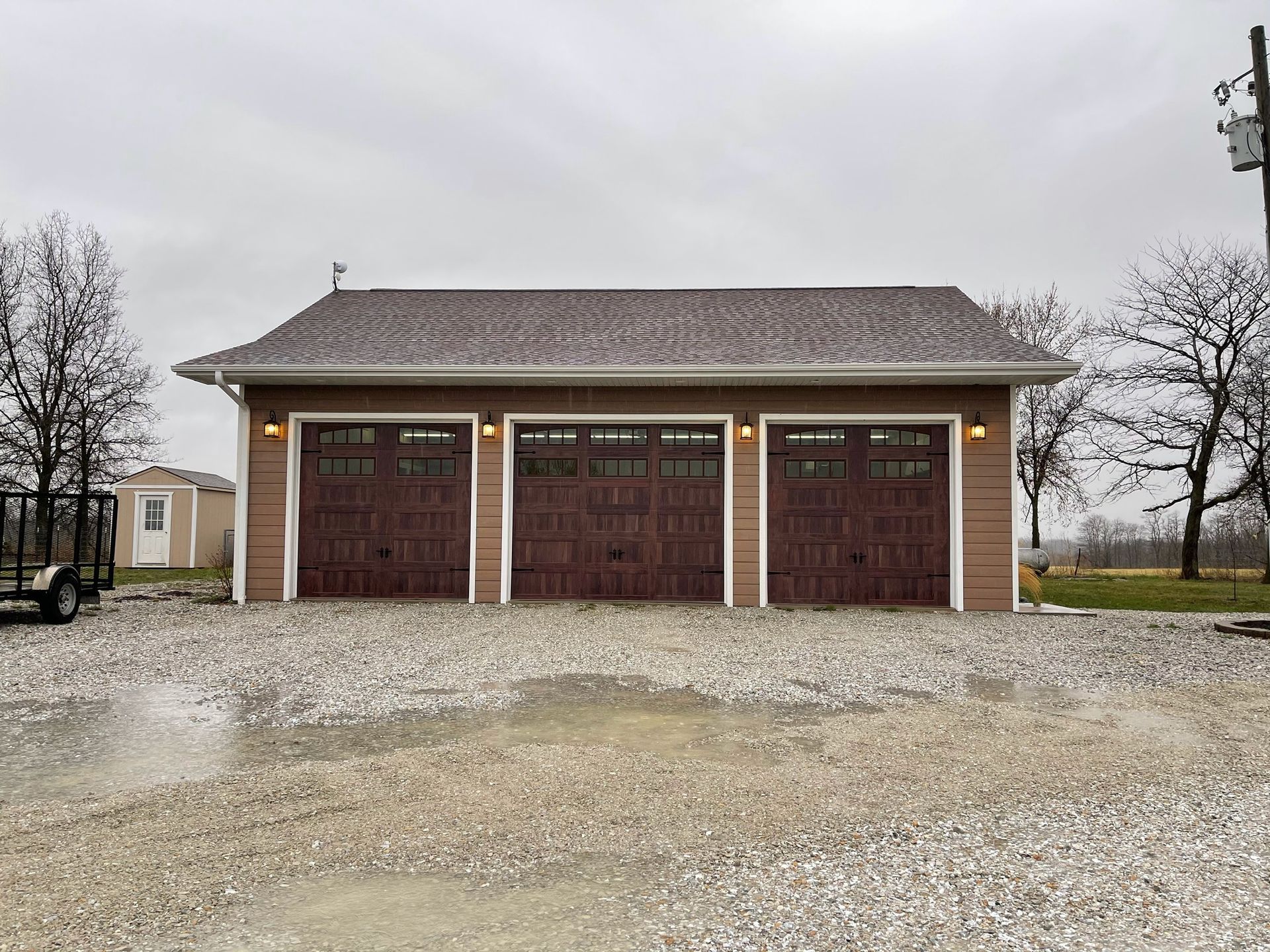 Small brown garage with three doors and a gravel driveway under a cloudy sky
