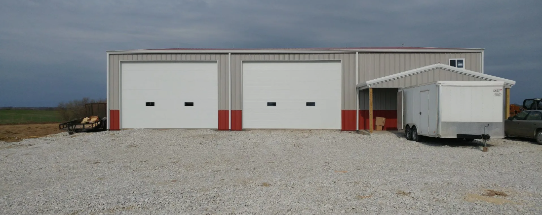 Two-bay garage with red accents, a small trailer, and gravel lot under a cloudy sky.