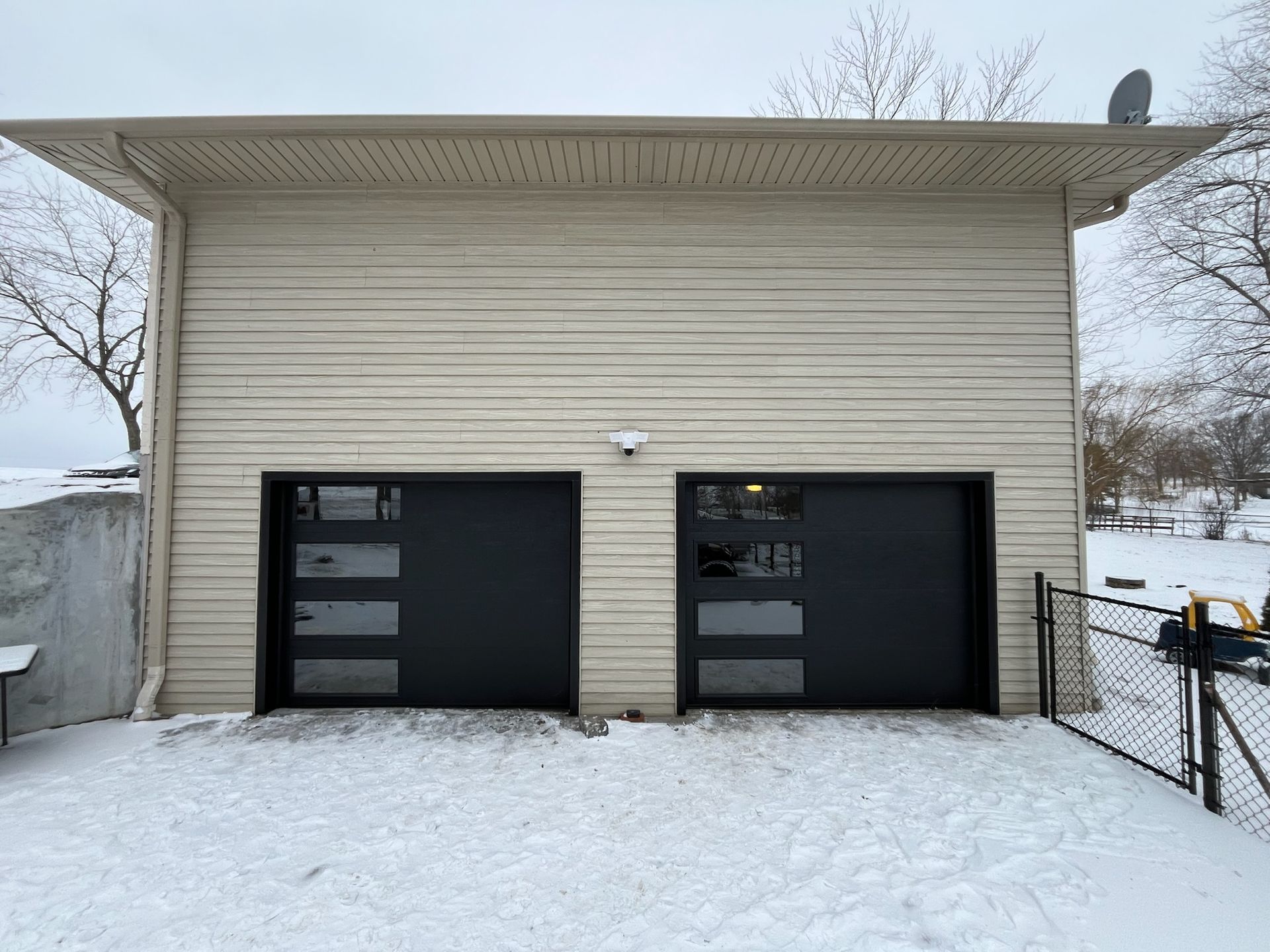 Two-car garage with black doors and glass panels. Beige siding. Snow on ground.