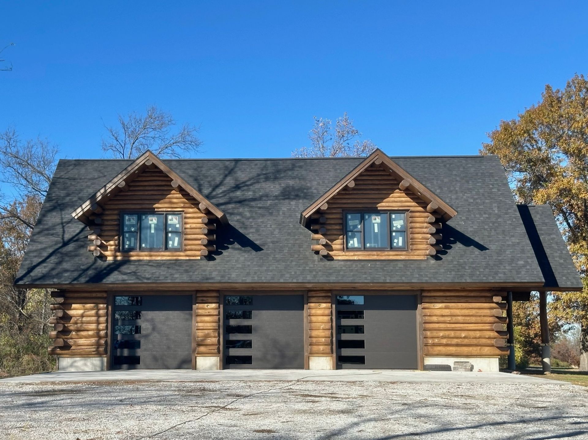 Rustic log cabin with two garage doors, dormer windows, and a snowy driveway under a clear blue sky