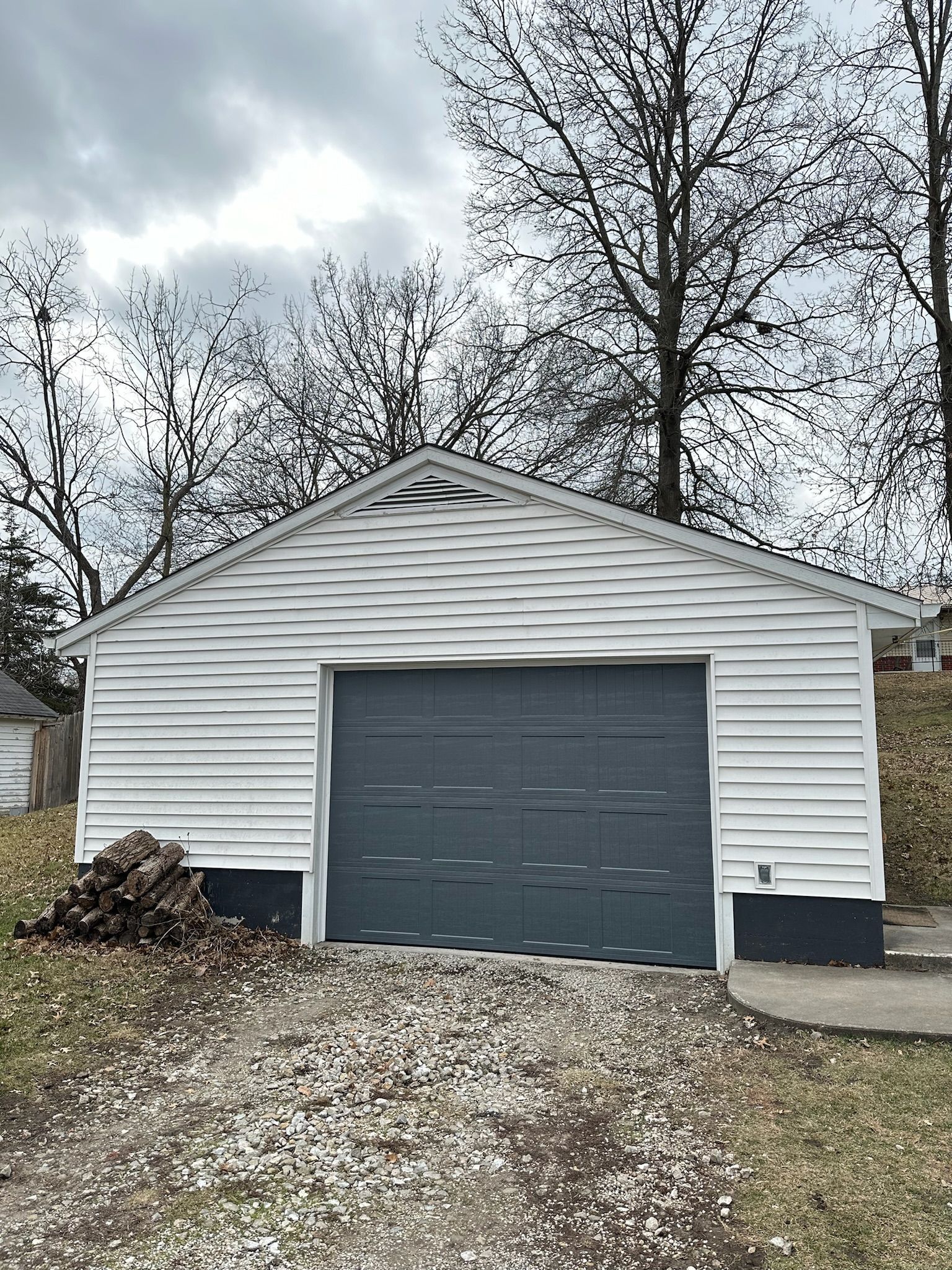 White-sided garage with a gray door, gravel driveway, and leafless trees against a cloudy sky.