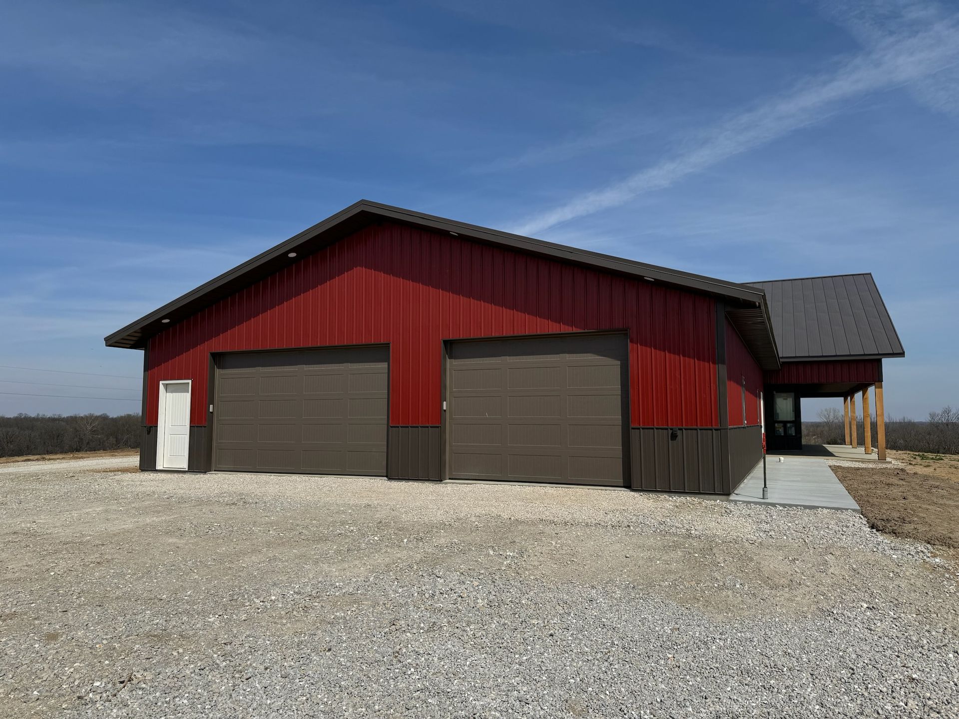 Red and brown metal building with two garage doors, gravel driveway, and a porch under a blue sky.
