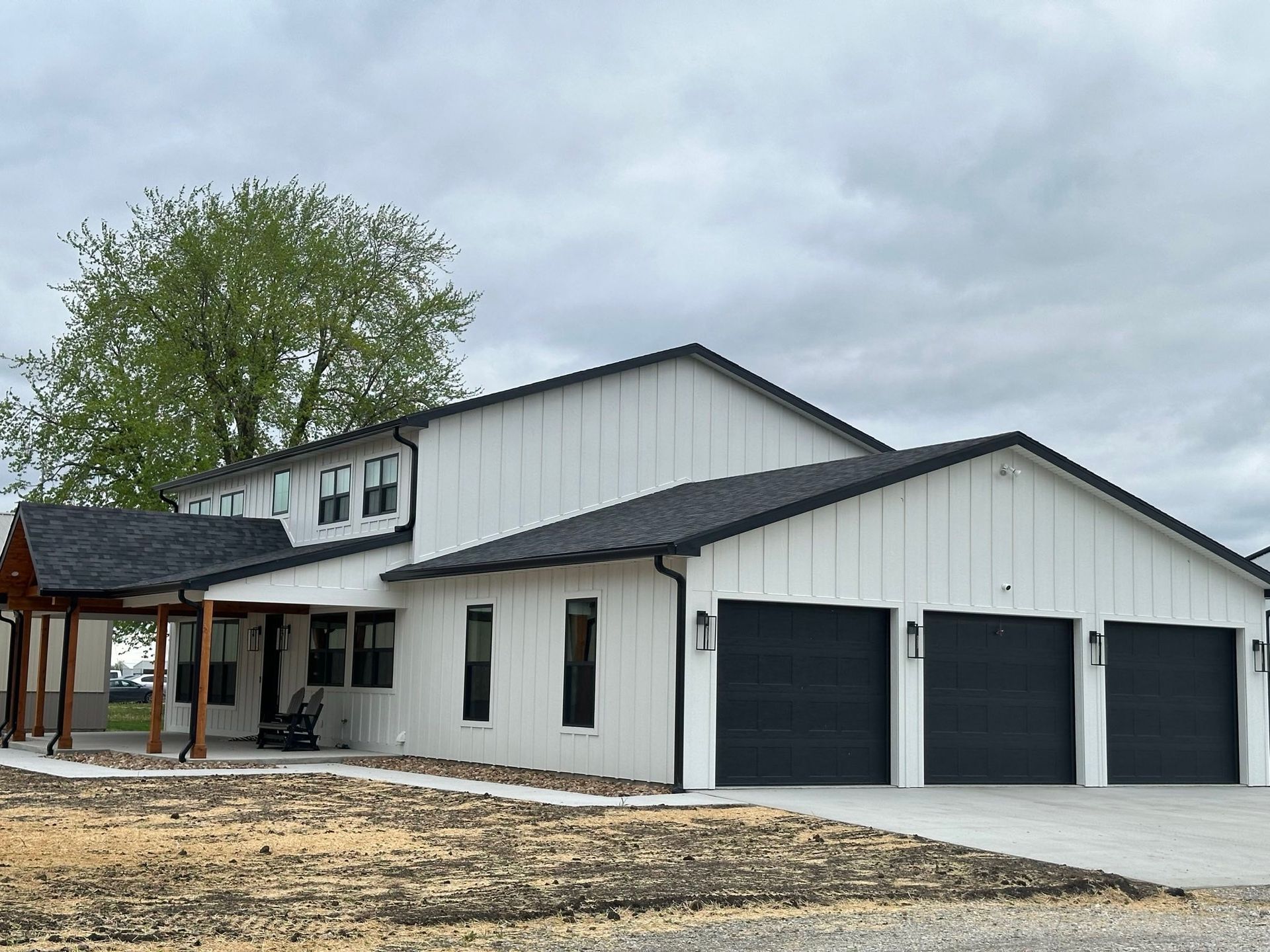 White house with black garage doors and covered porch, set on a dirt lot under a cloudy sky