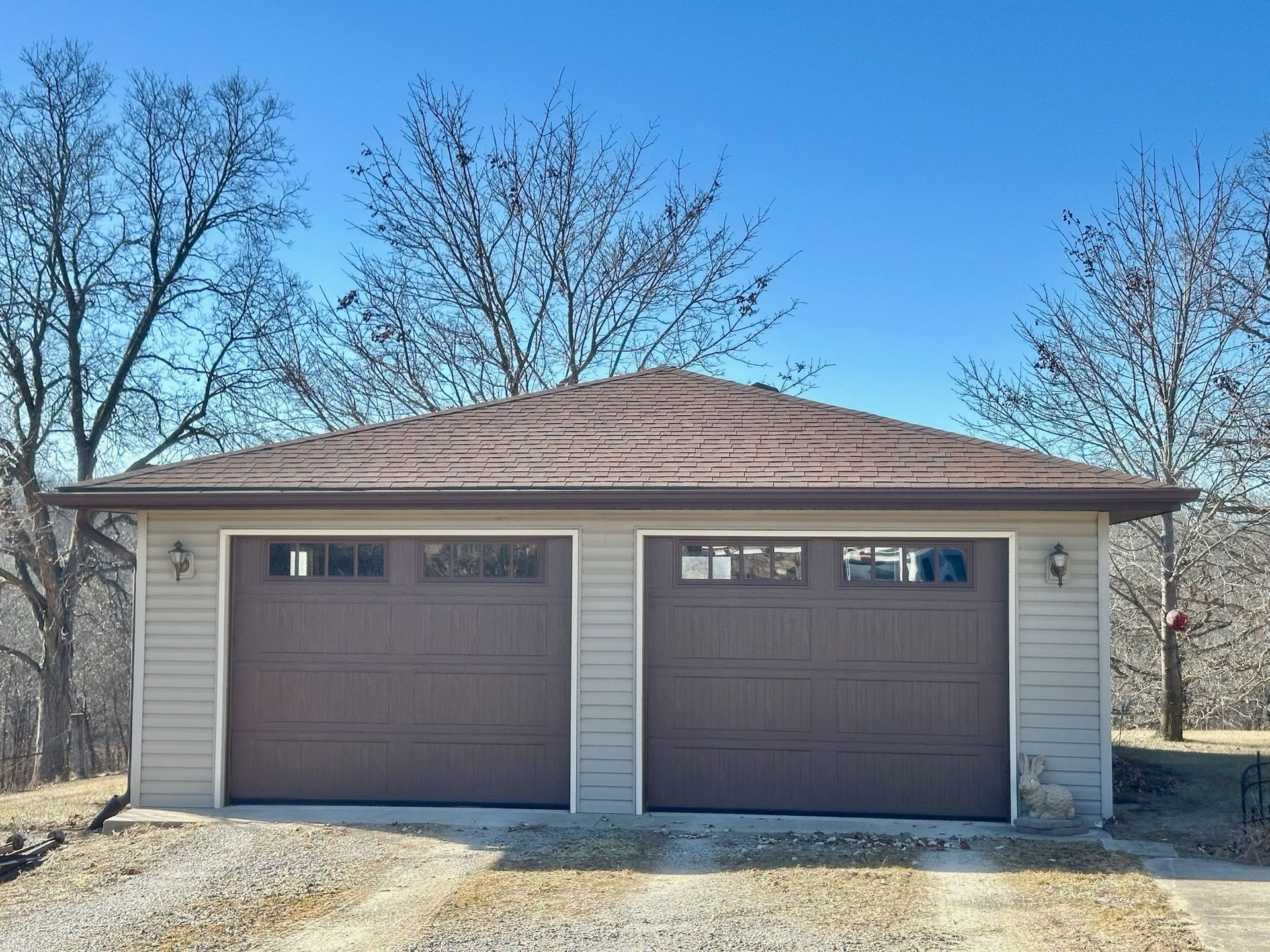 Two-car garage with brown doors, tan siding, and brown roof under a clear, blue sky.