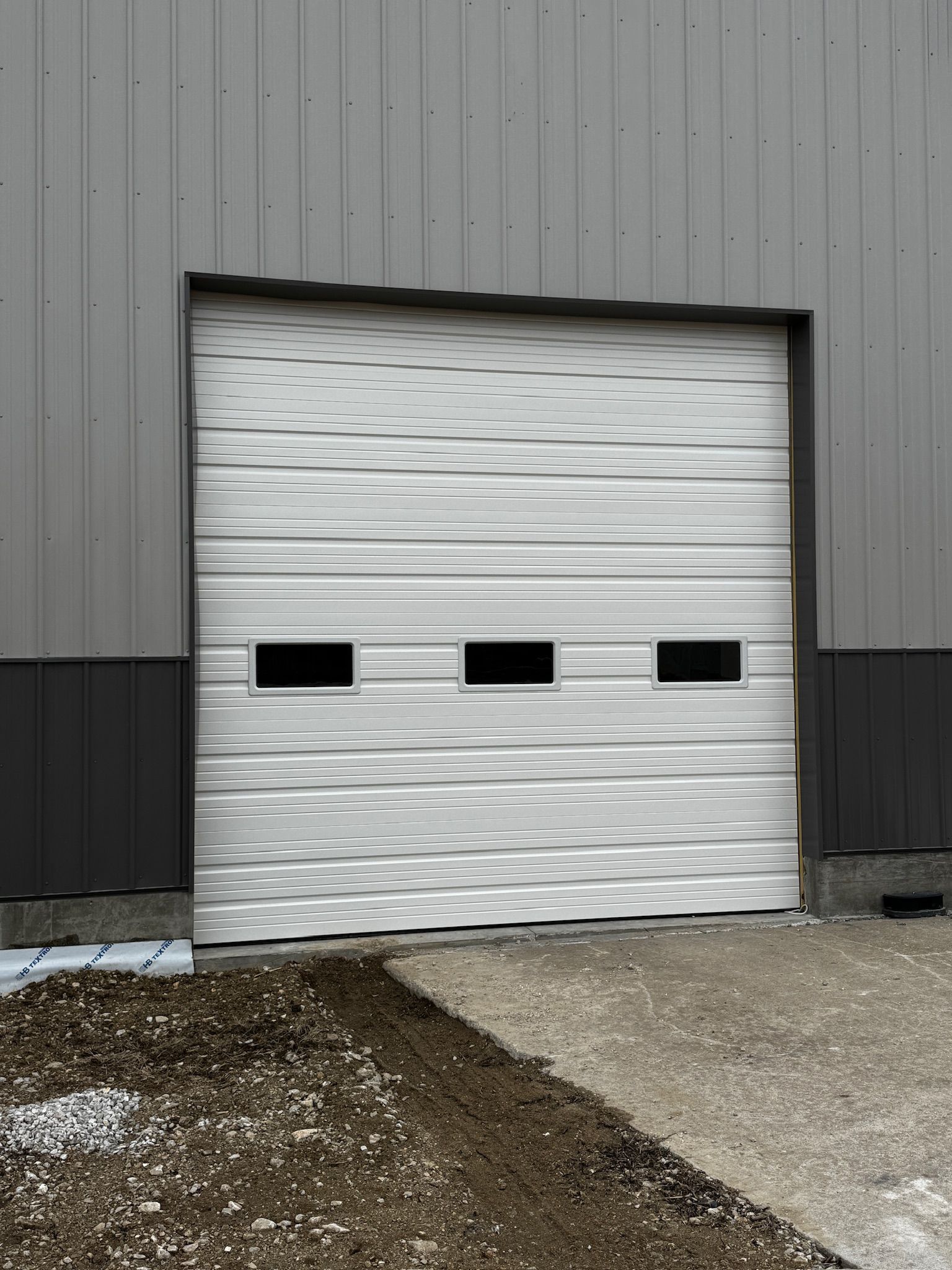 White metal roll-up garage door with three windows, gray building exterior, gravel ground.
