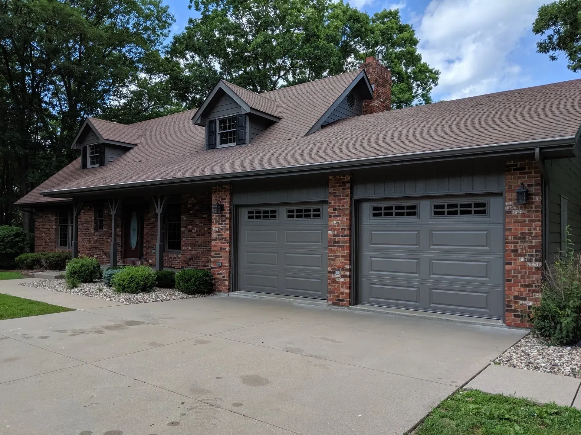Brick house with gray garage doors, dormers, and a brown roof. Driveway and landscaping in front.